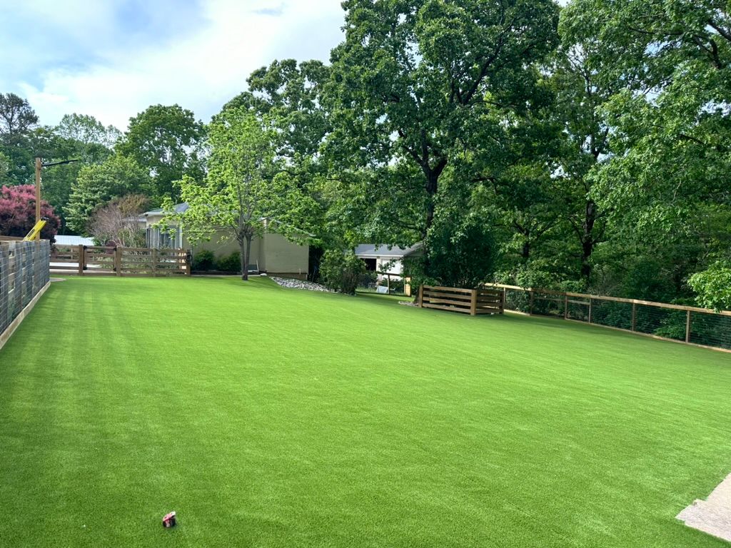Lush green lawn surrounded by a wooden fence, trees, and a small building under a blue sky.