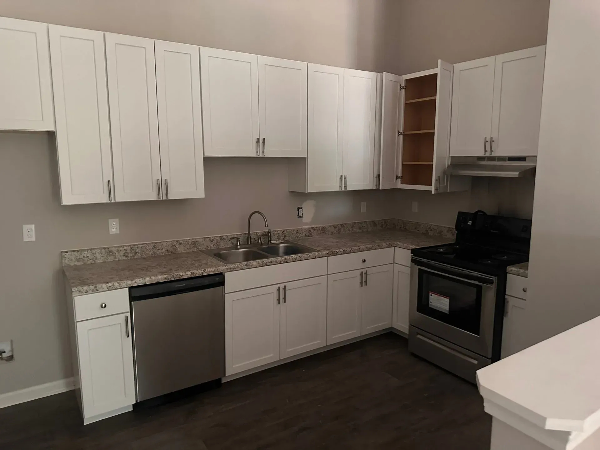 Empty kitchen with white cabinets, granite countertops, stainless dishwasher, black stove, and dark wood floor.