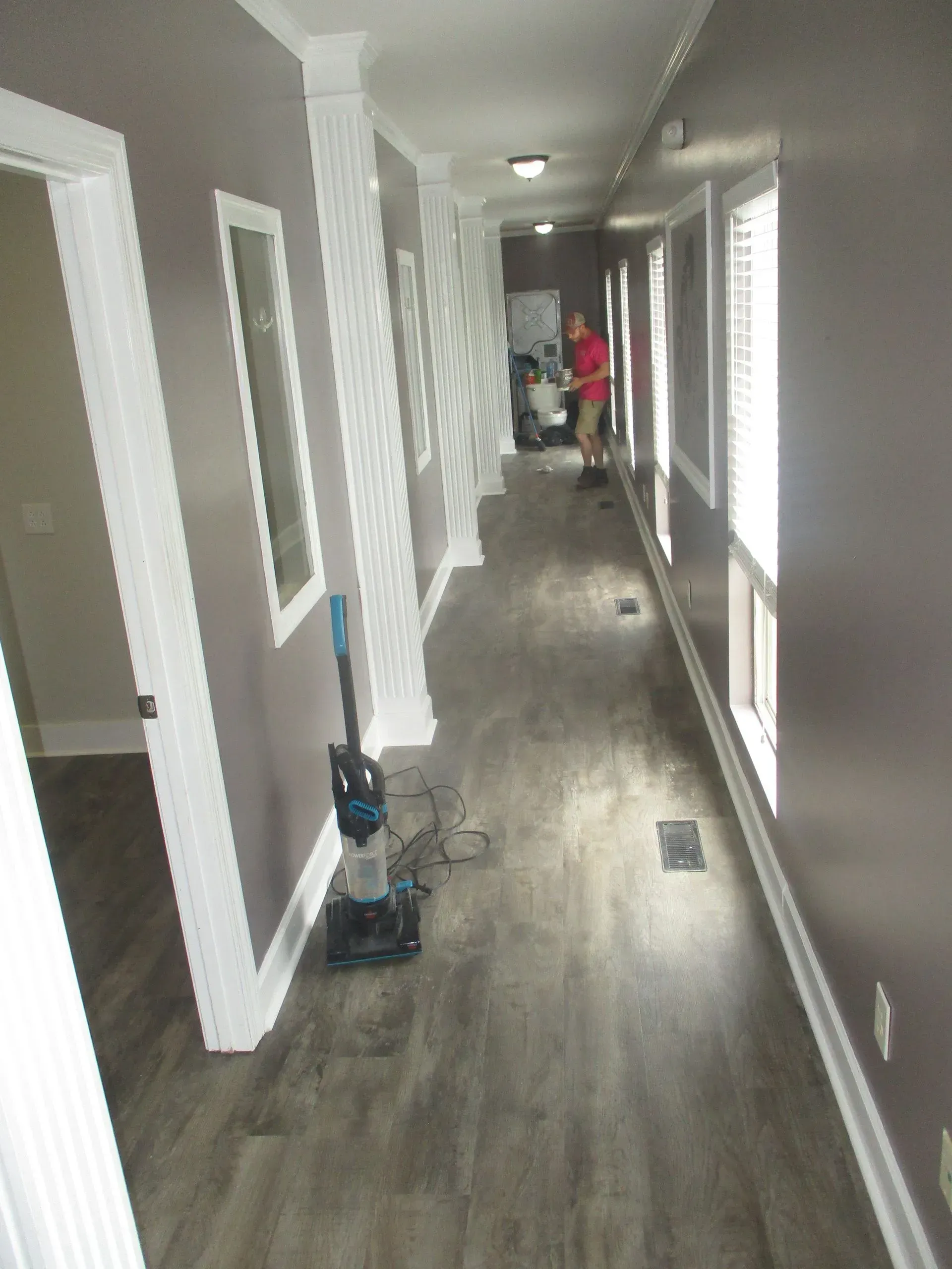 Long hallway with gray wood floors, white doors, and a person cleaning in the distance.