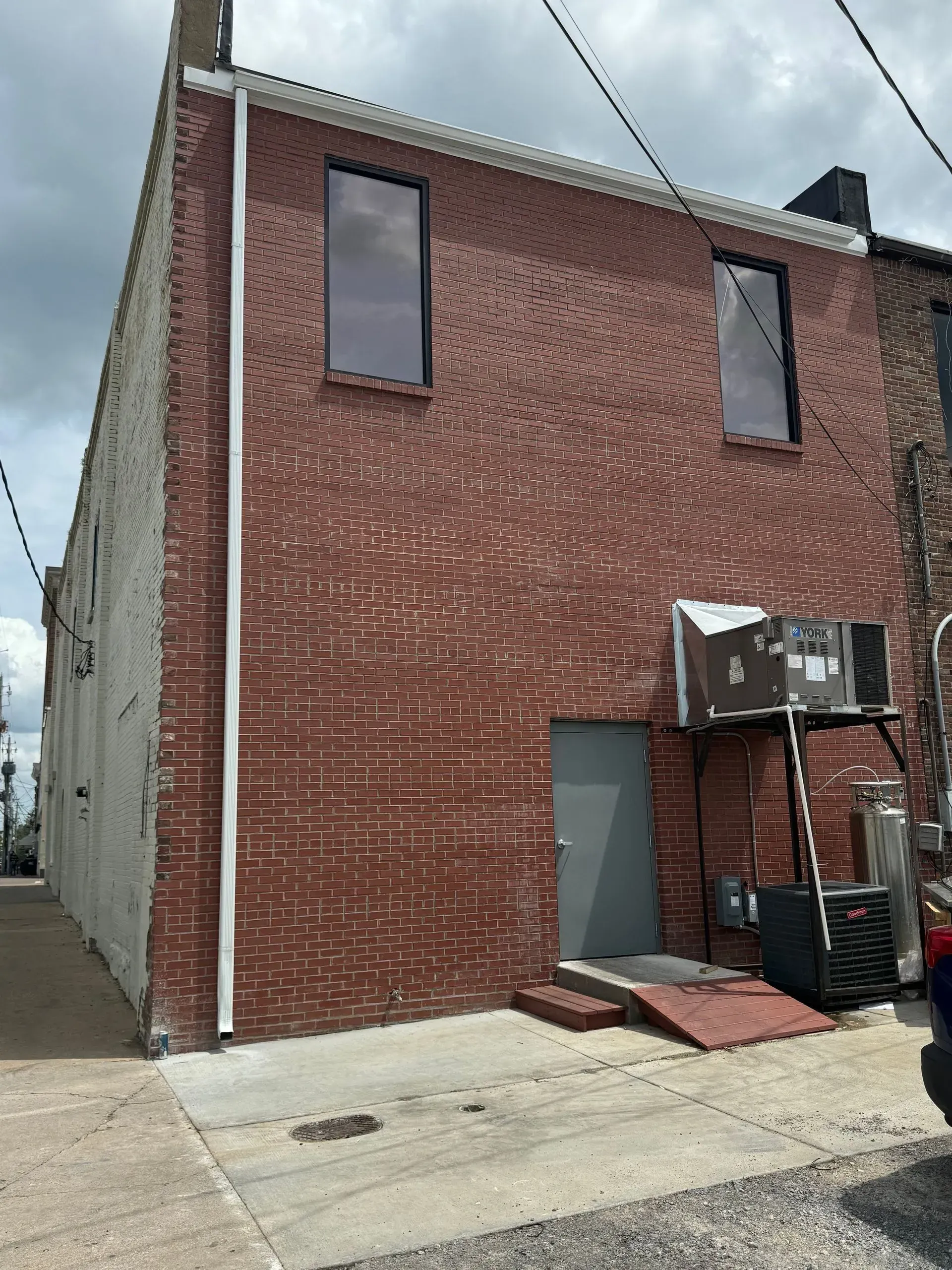 Brick building exterior with gray side wall, narrow windows, and utility units above a red rear door.