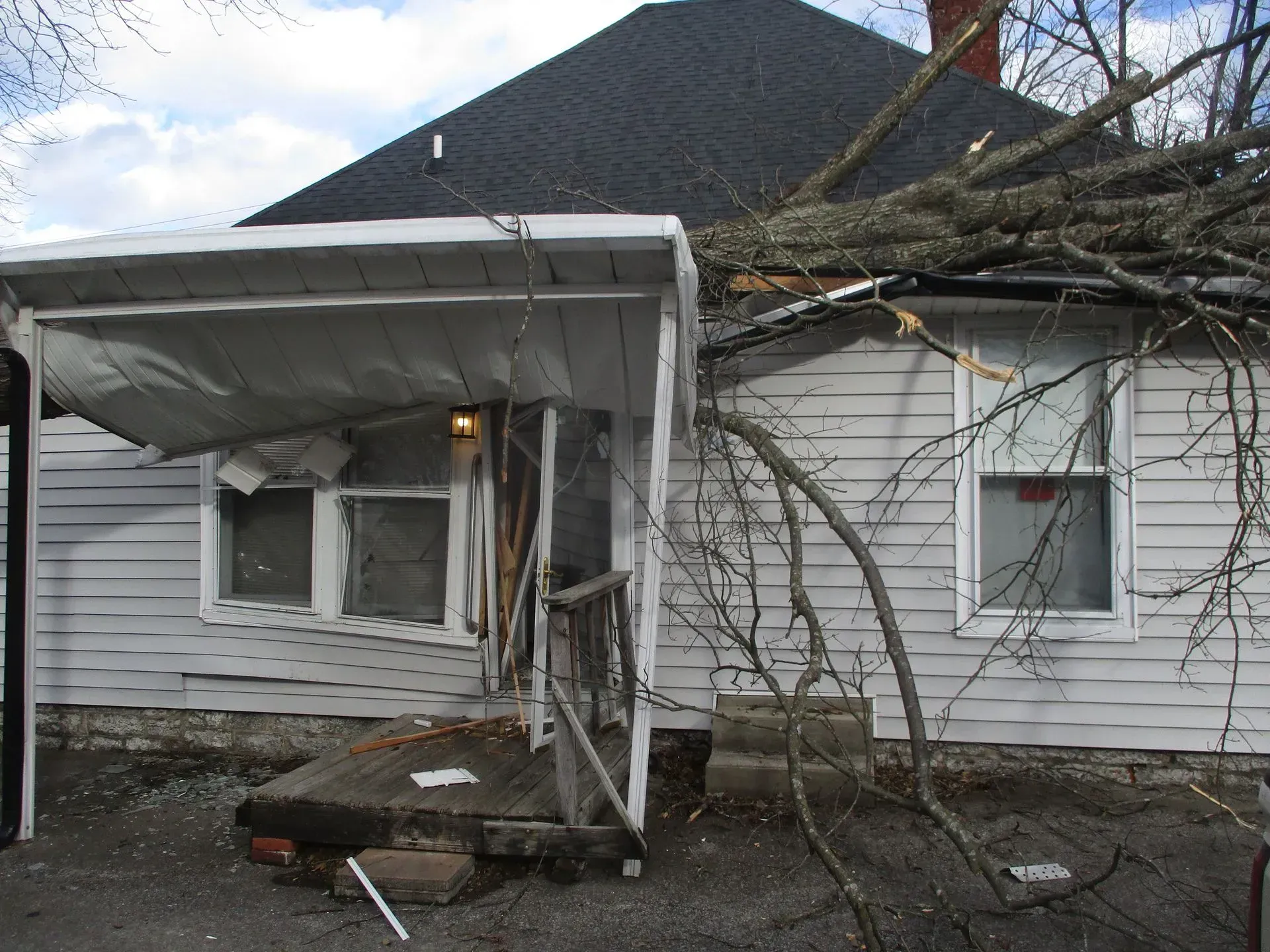 Damaged white house with collapsed porch roof and fallen tree branches after a storm