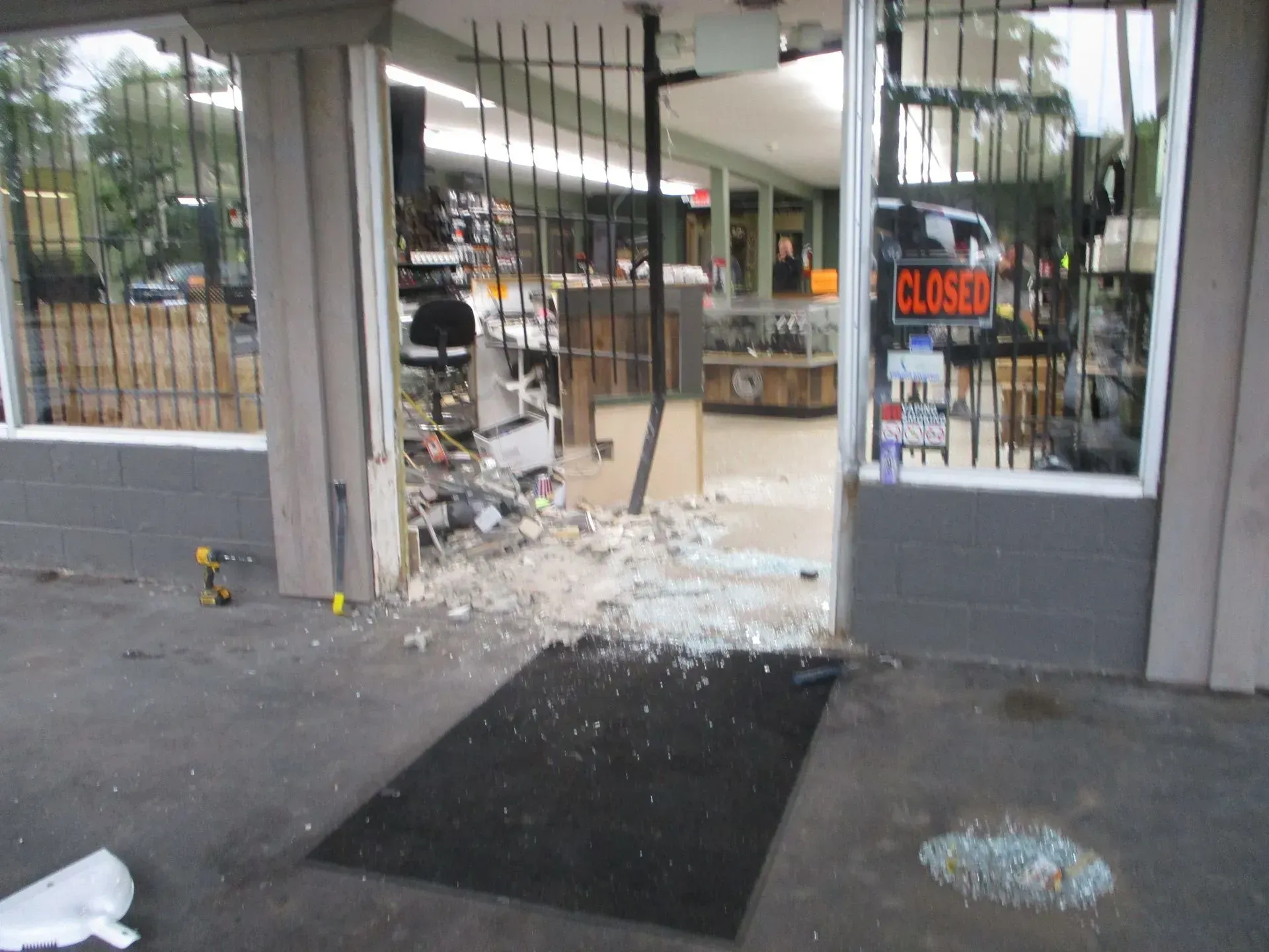 Damaged storefront with shattered glass, broken door frame, and debris on the sidewalk; closed sign visible inside