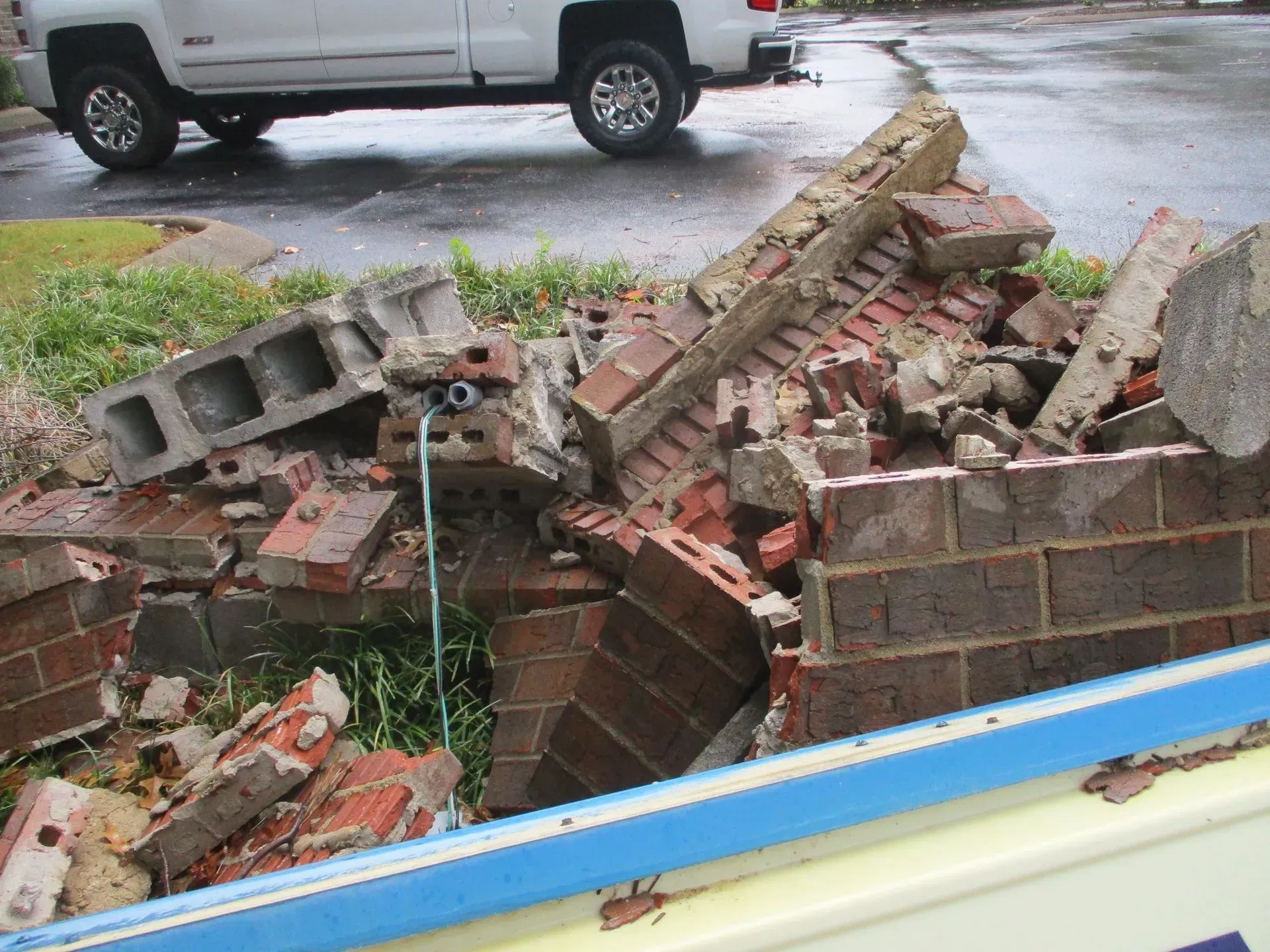Collapsed brick wall and rubble beside a driveway, with a truck in the background.