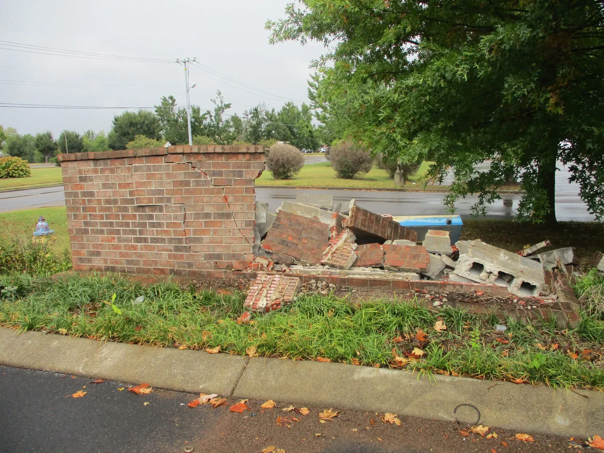 Collapsed brick wall along a roadside curb with scattered debris and flowers nearby