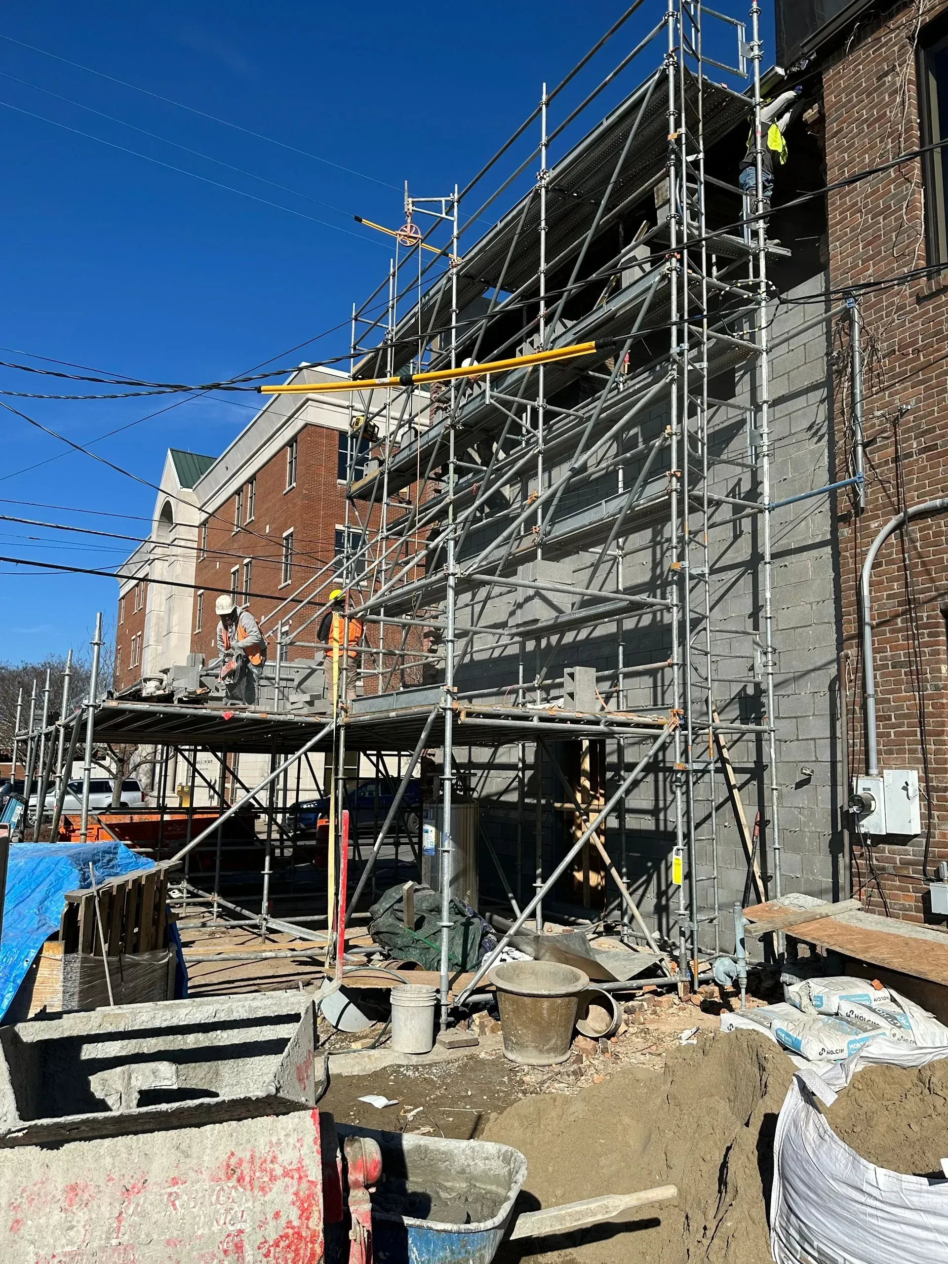 Scaffolding on a brick building under construction beside a sidewalk and piled dirt, under a clear blue sky