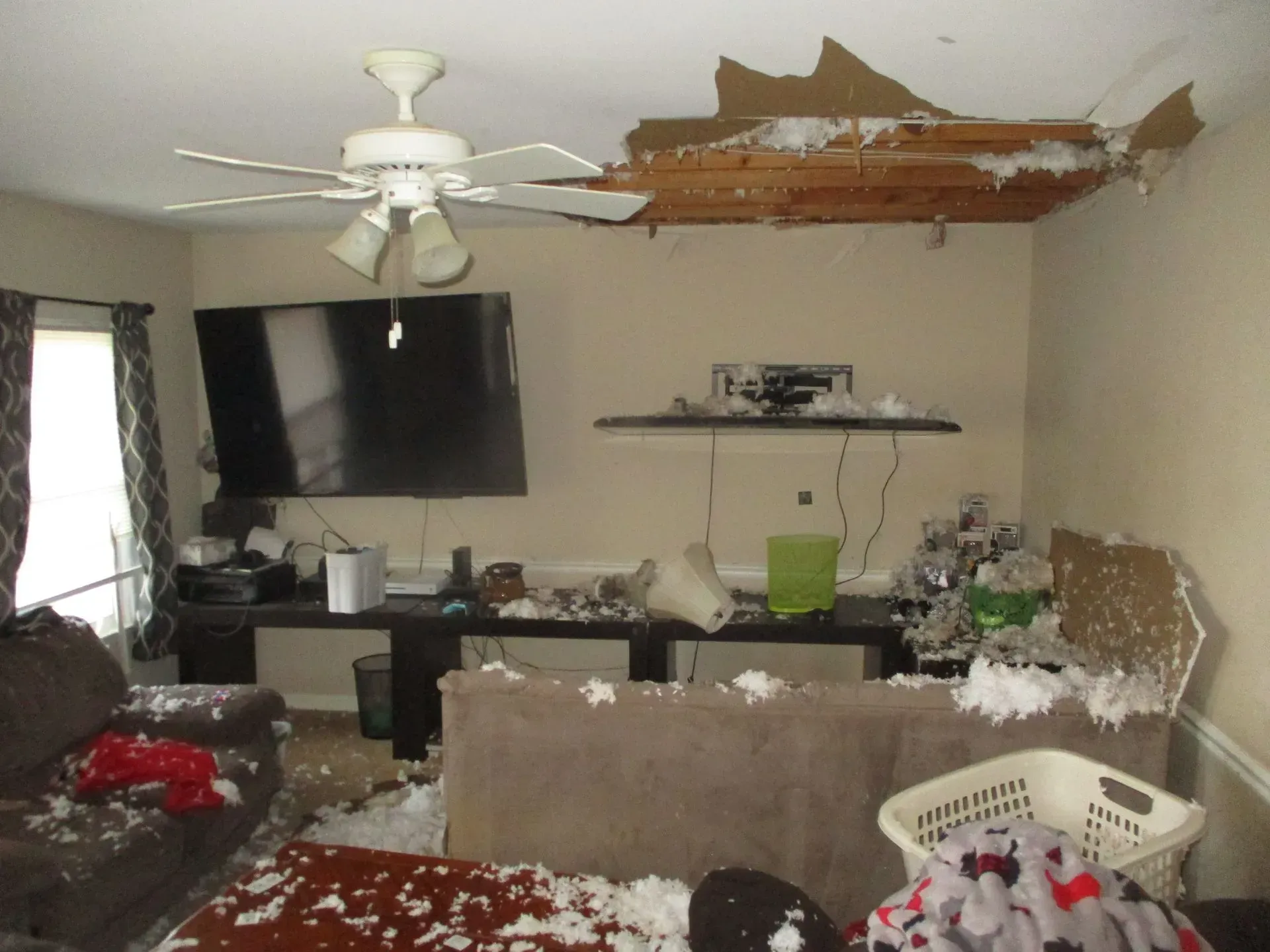 Living room with damaged ceiling, exposed wood, and scattered debris around a couch and TV