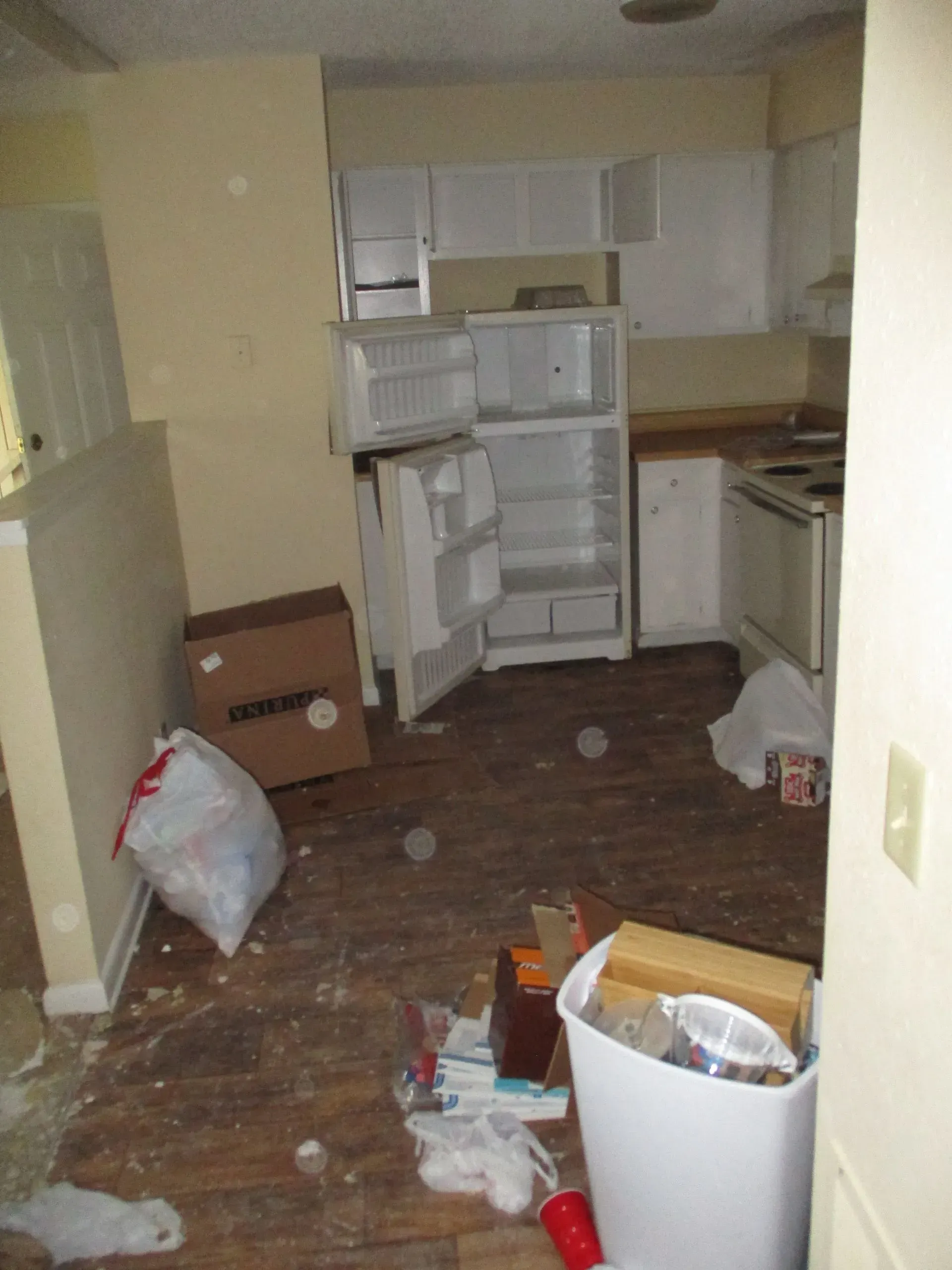 Cluttered kitchen with open refrigerator, scattered trash, and boxes on the floor