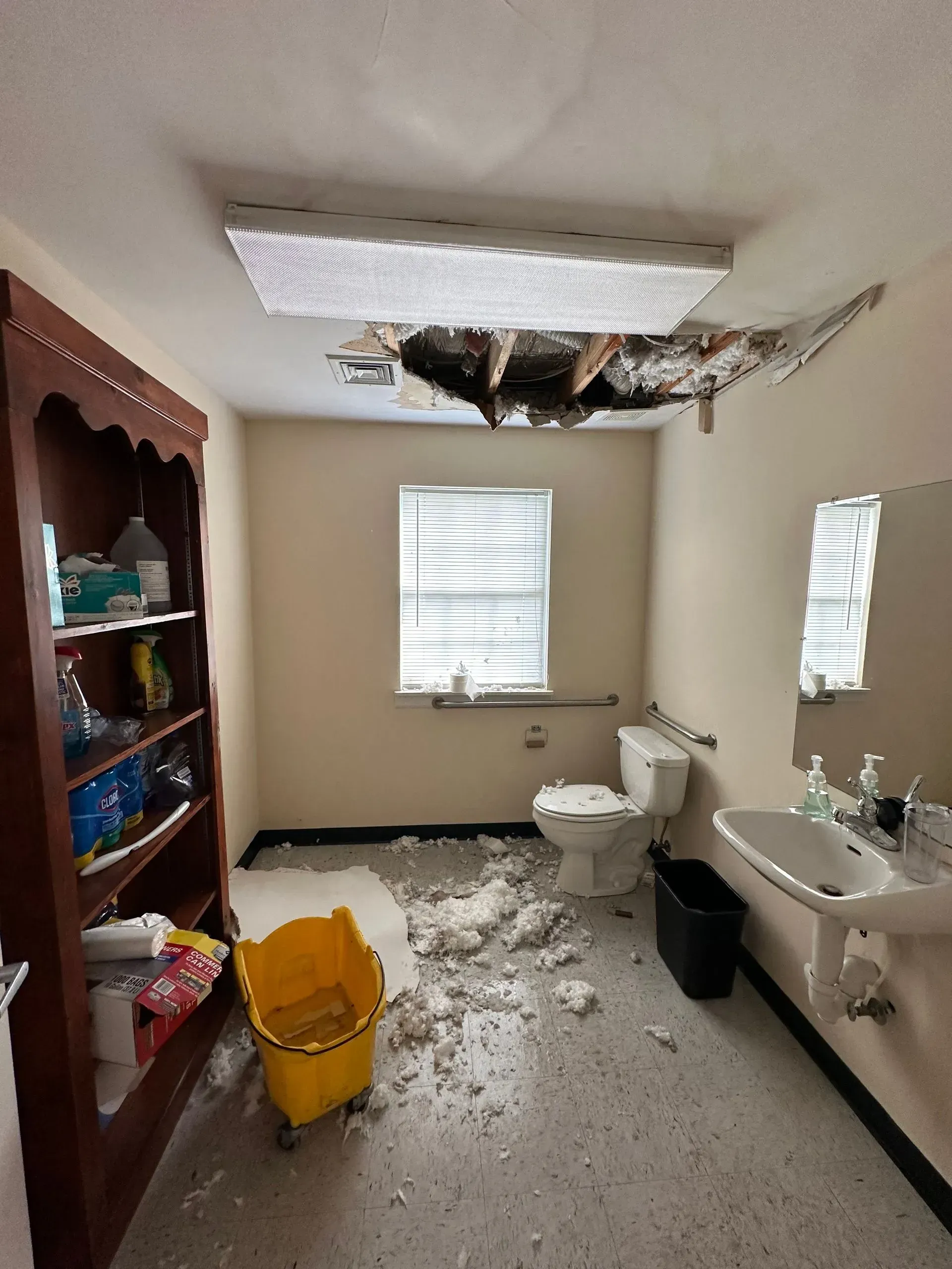 Damaged bathroom with hole in the ceiling, debris on floor, sink, toilet, and yellow bucket