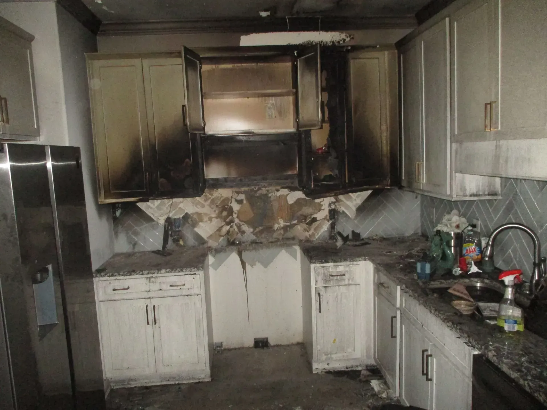 Fire-damaged kitchen with charred cabinets, melted appliances, and debris on the counters and floor
