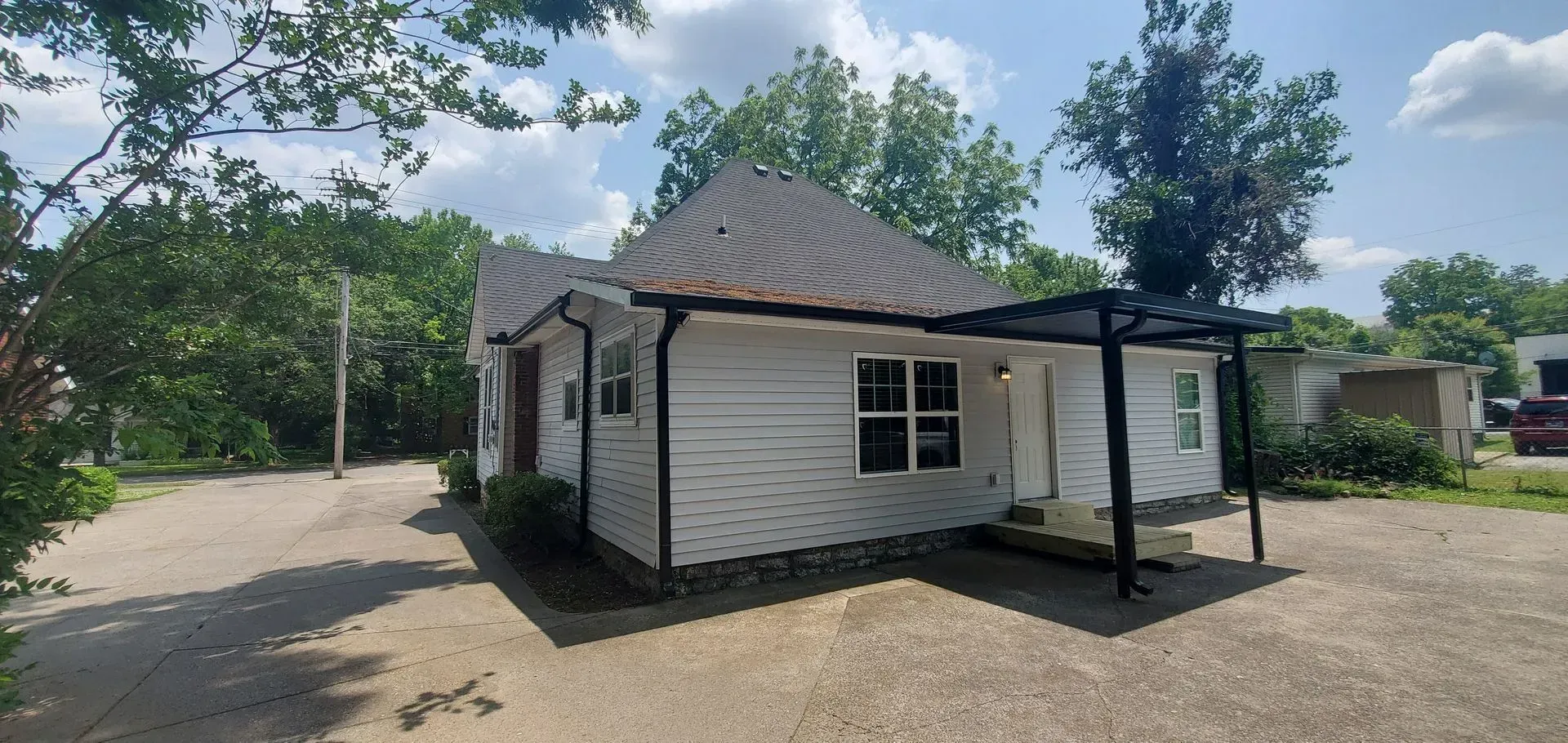 Small white house with a wavy roof and black porch frame beside a paved lot and trees