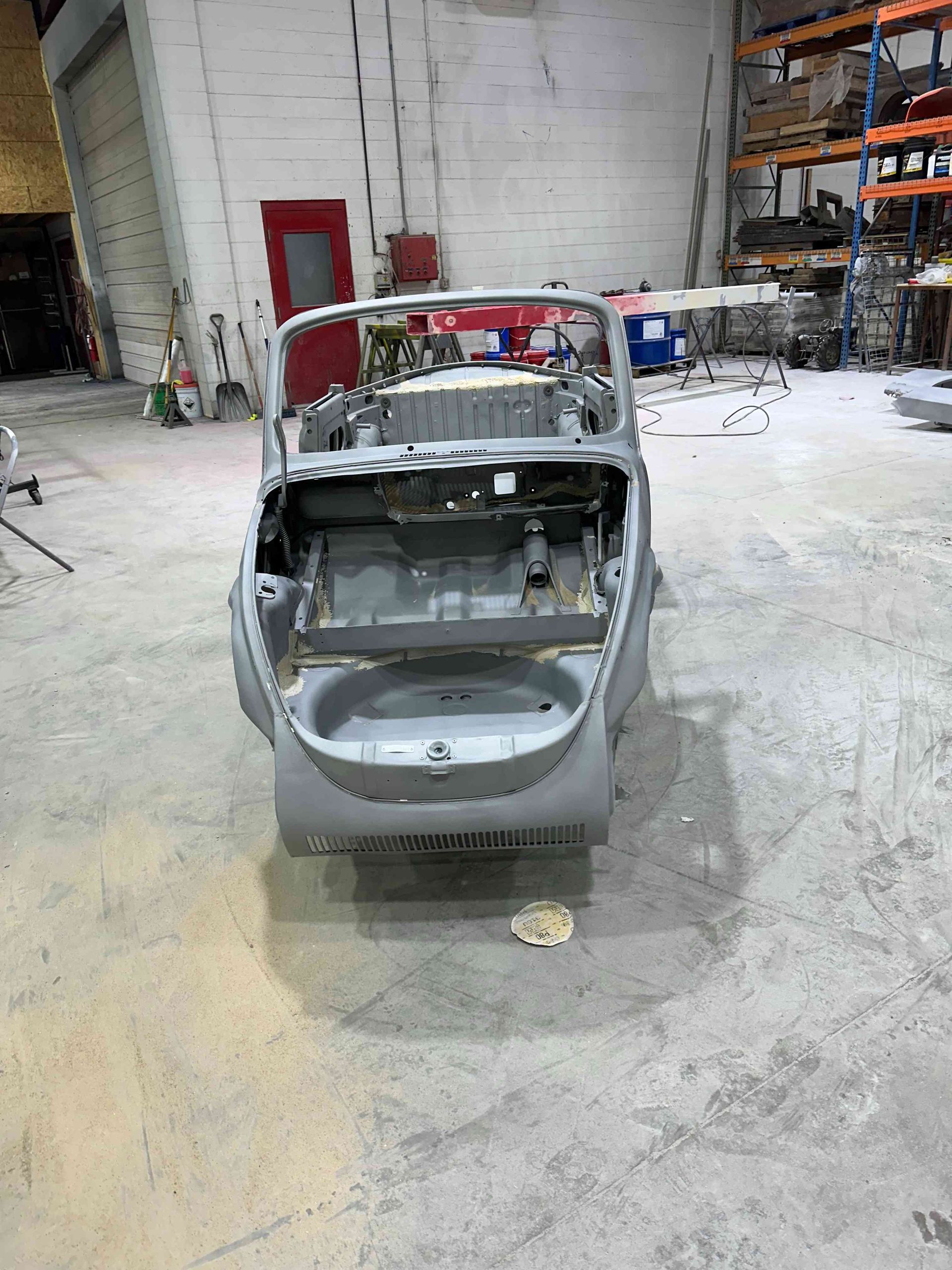 Gray car shell in an auto shop, rear hatch open and interior stripped, with shelves and equipment in background