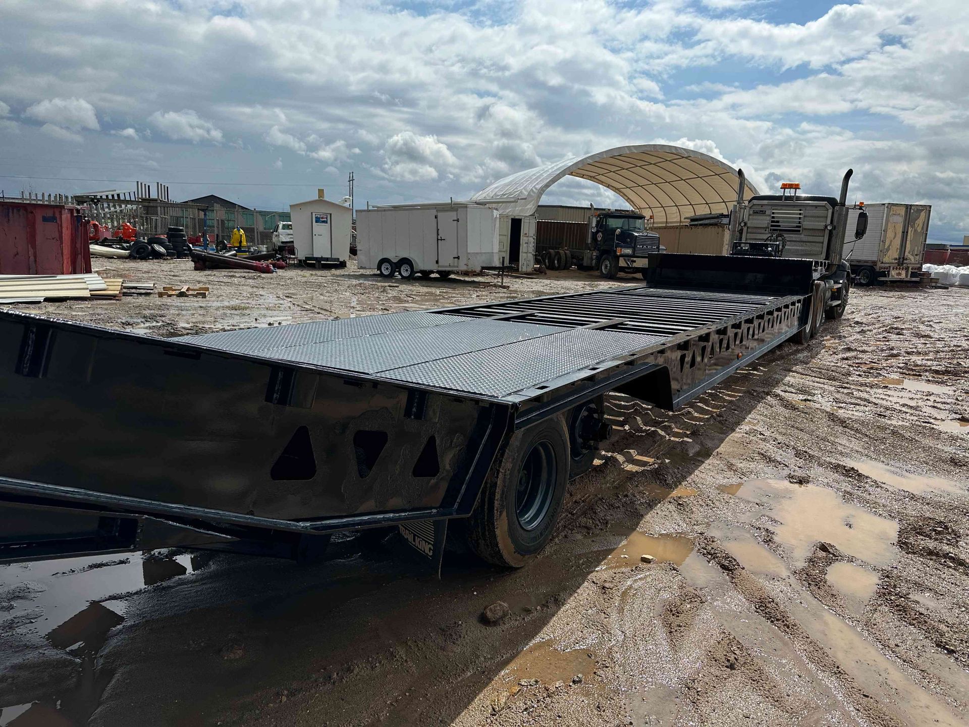 Flatbed trailer in a muddy lot under cloudy sky, with a white truck and canopy in the background.