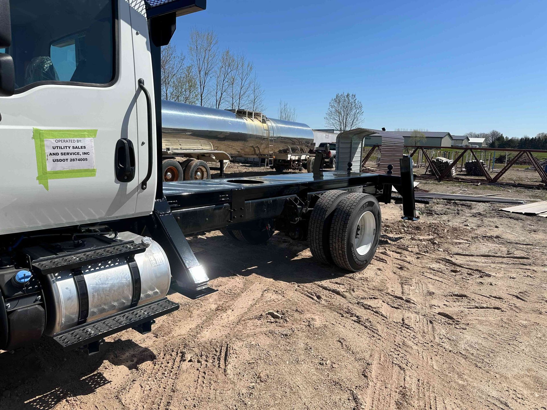 White truck chassis parked on dirt lot under clear blue sky, with lumber and equipment in the background