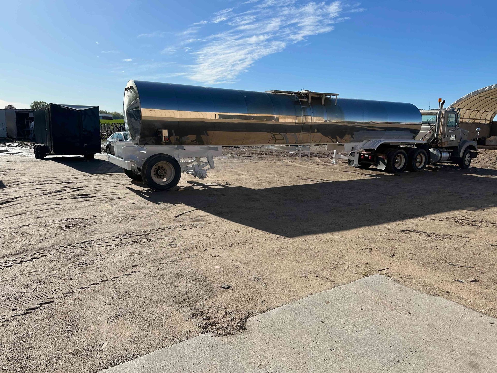 Stainless steel tanker trailer parked on a dirt lot under a blue sky.
