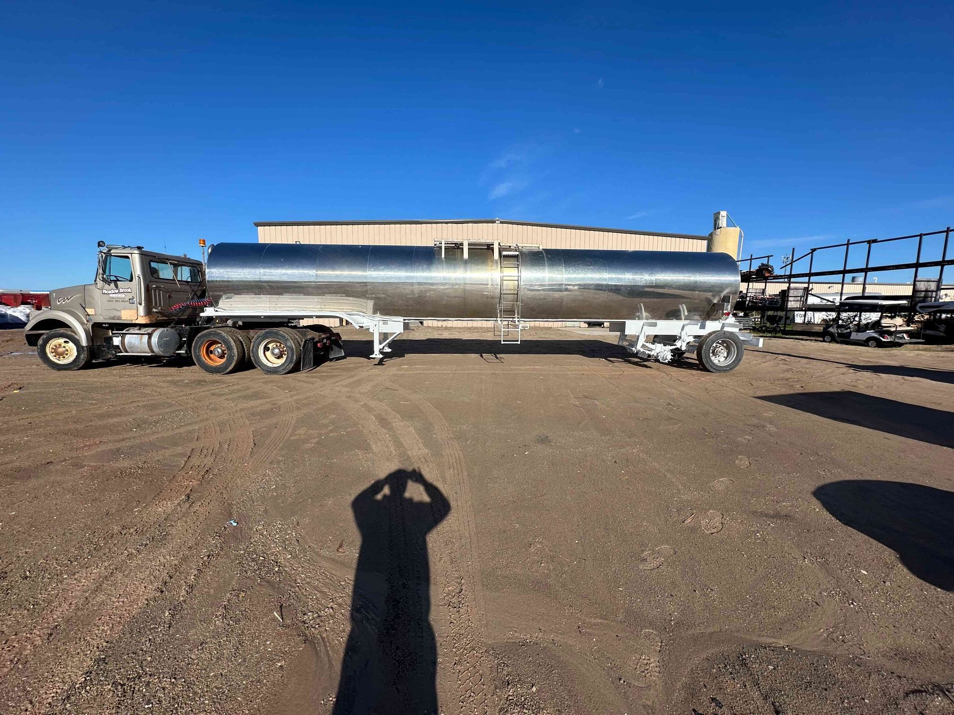 Semi-truck hauling a long silver cylindrical tank across a dirt lot under a clear blue sky