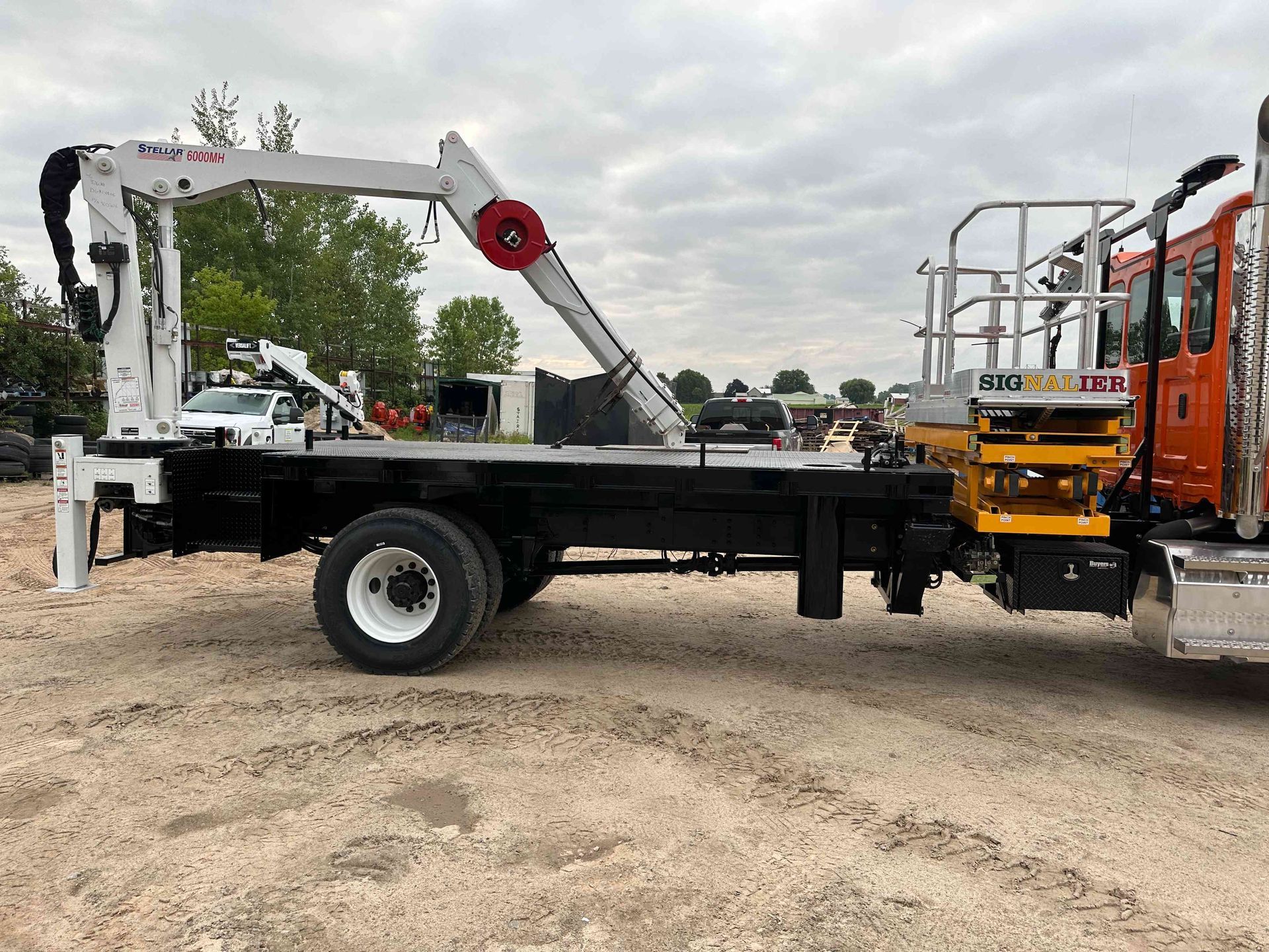 Tow truck lifting a white utility truck on a gravel lot, with a yellow truck cab beside it