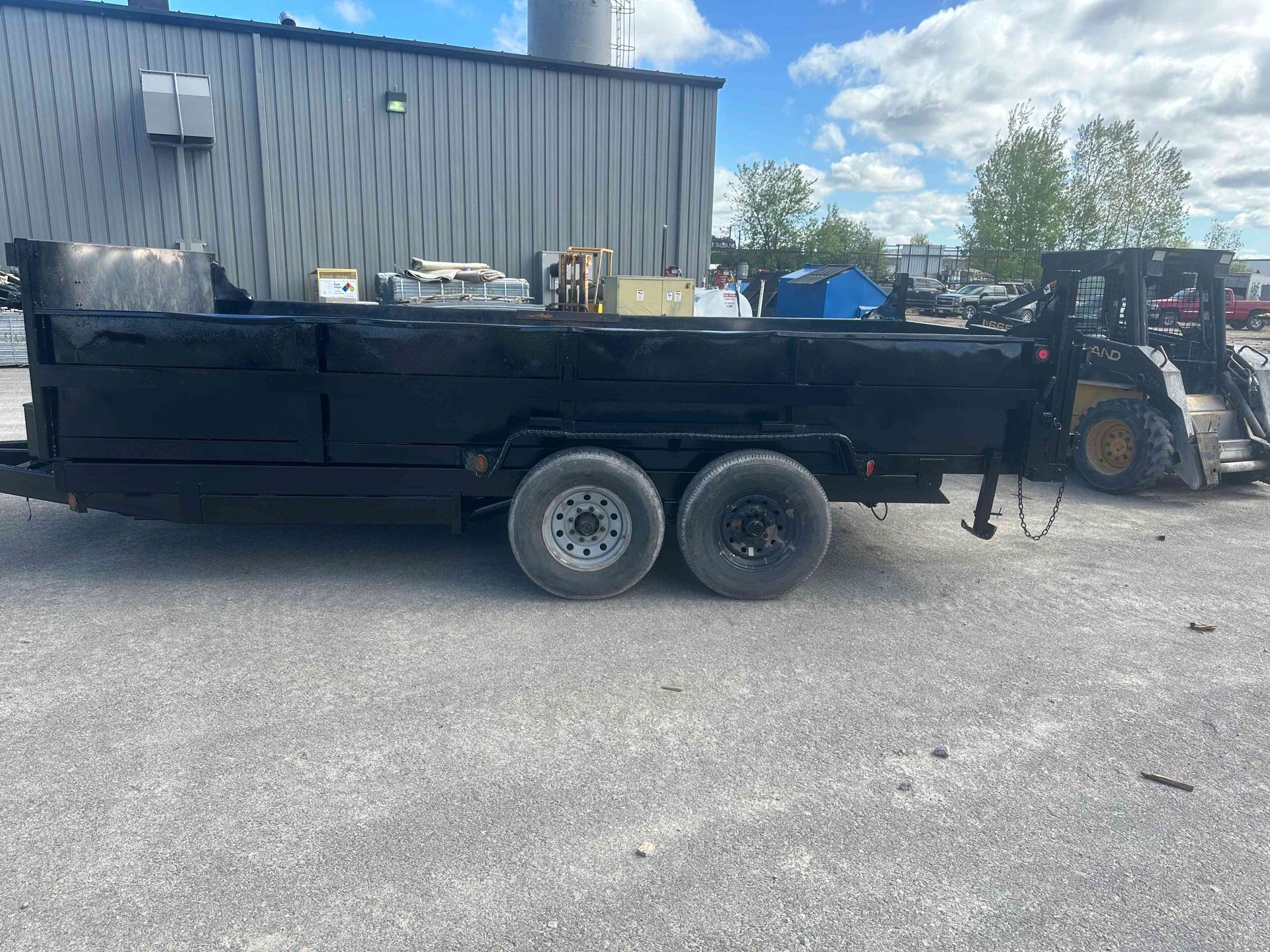 Black dump trailer with dual axles parked in an industrial lot beside a metal building.
