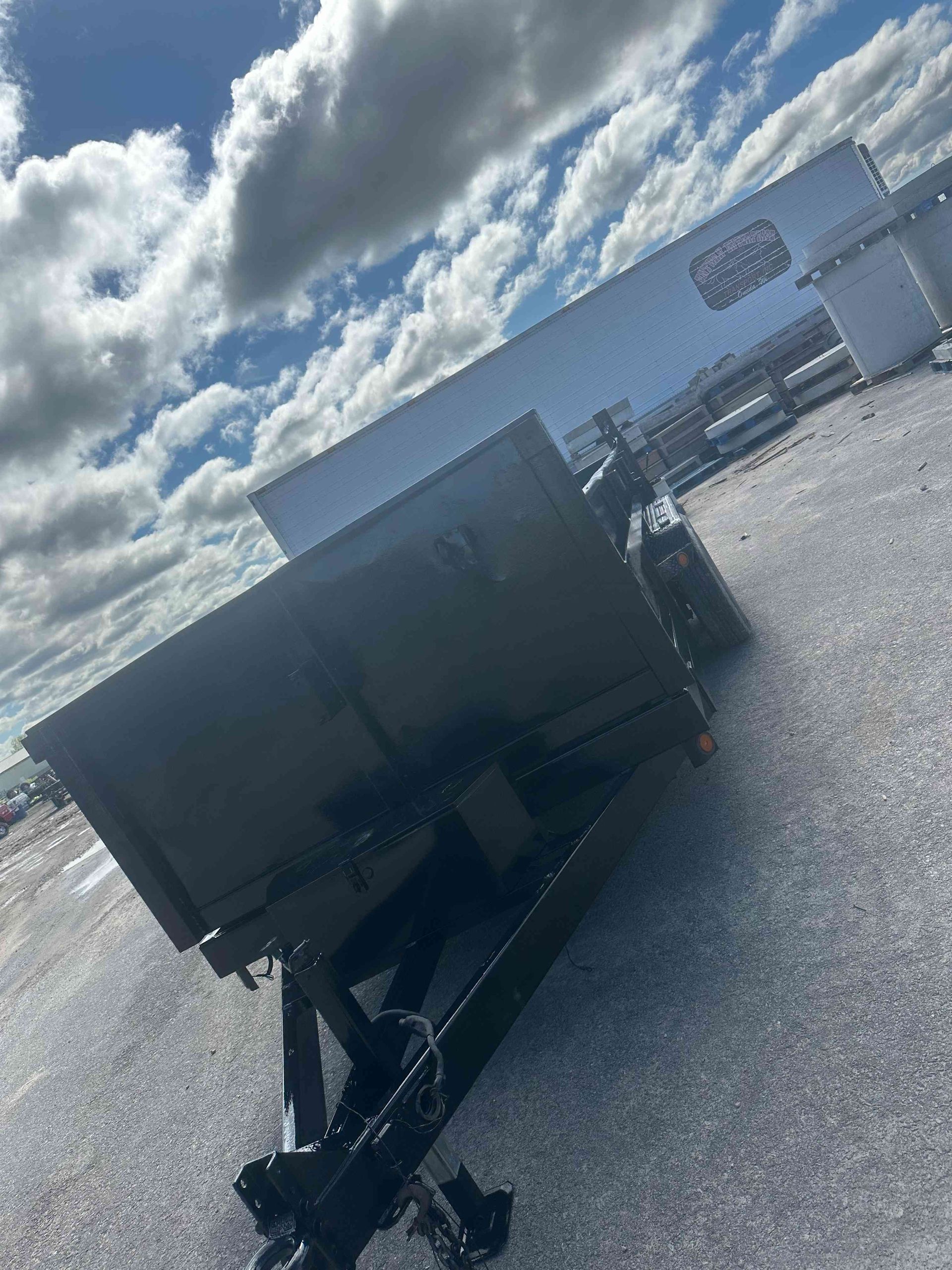 Black trailer on a wet lot under a cloudy sky beside a white industrial building.