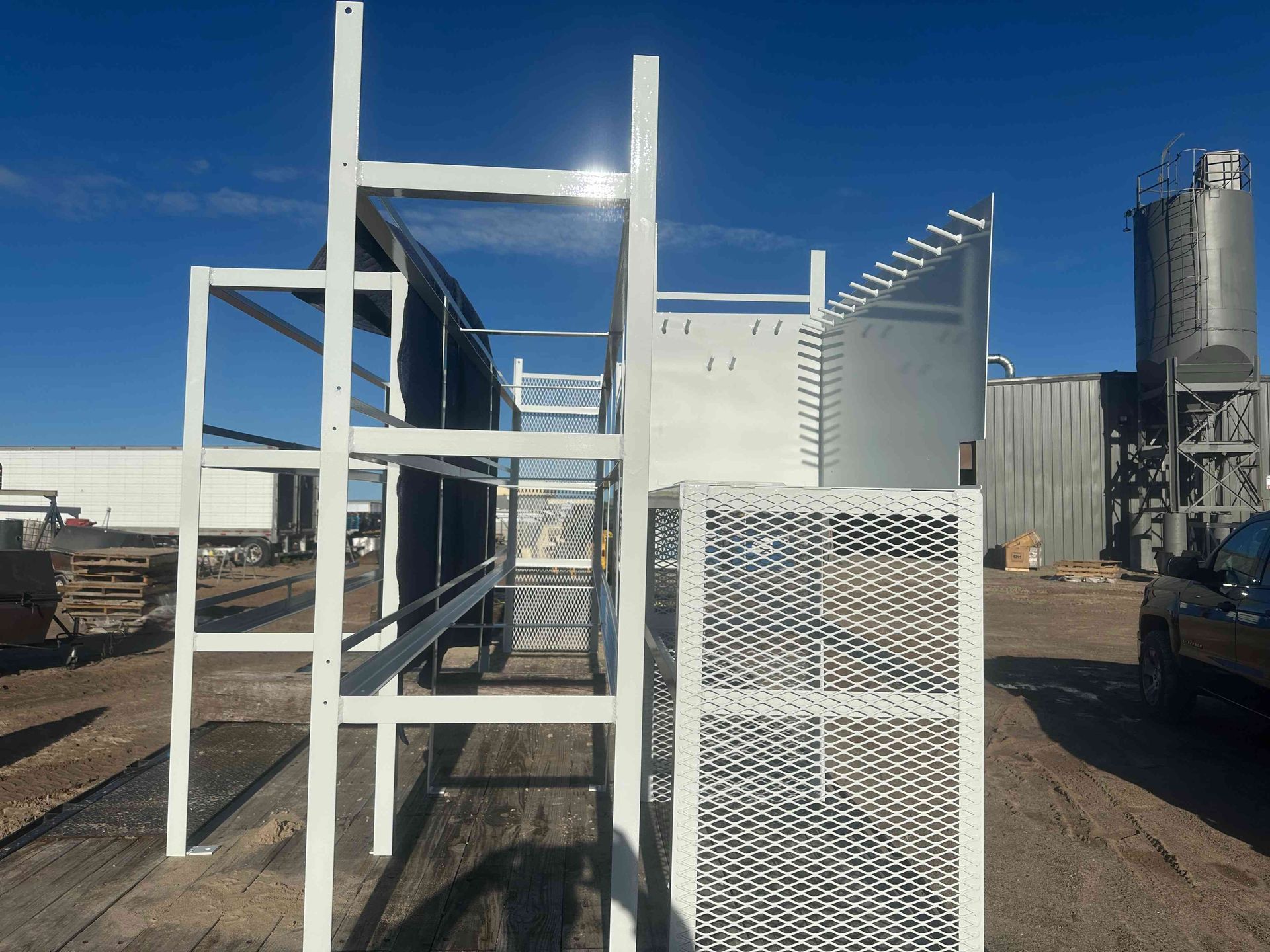 White metal industrial frame and mesh panels in a dirt yard beside a tall silo under a clear blue sky