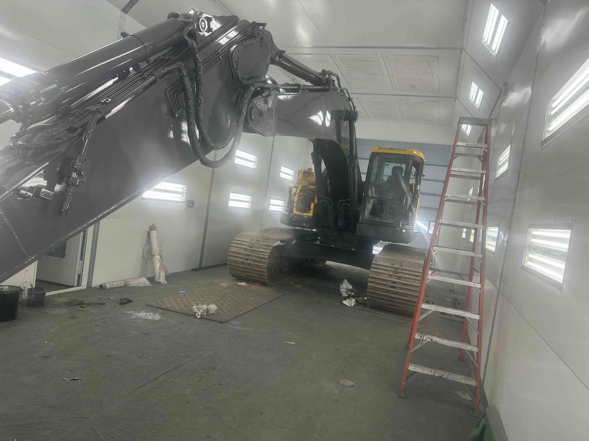 Excavator inside a metal enclosure, arm raised, with a red ladder leaning against the wall.