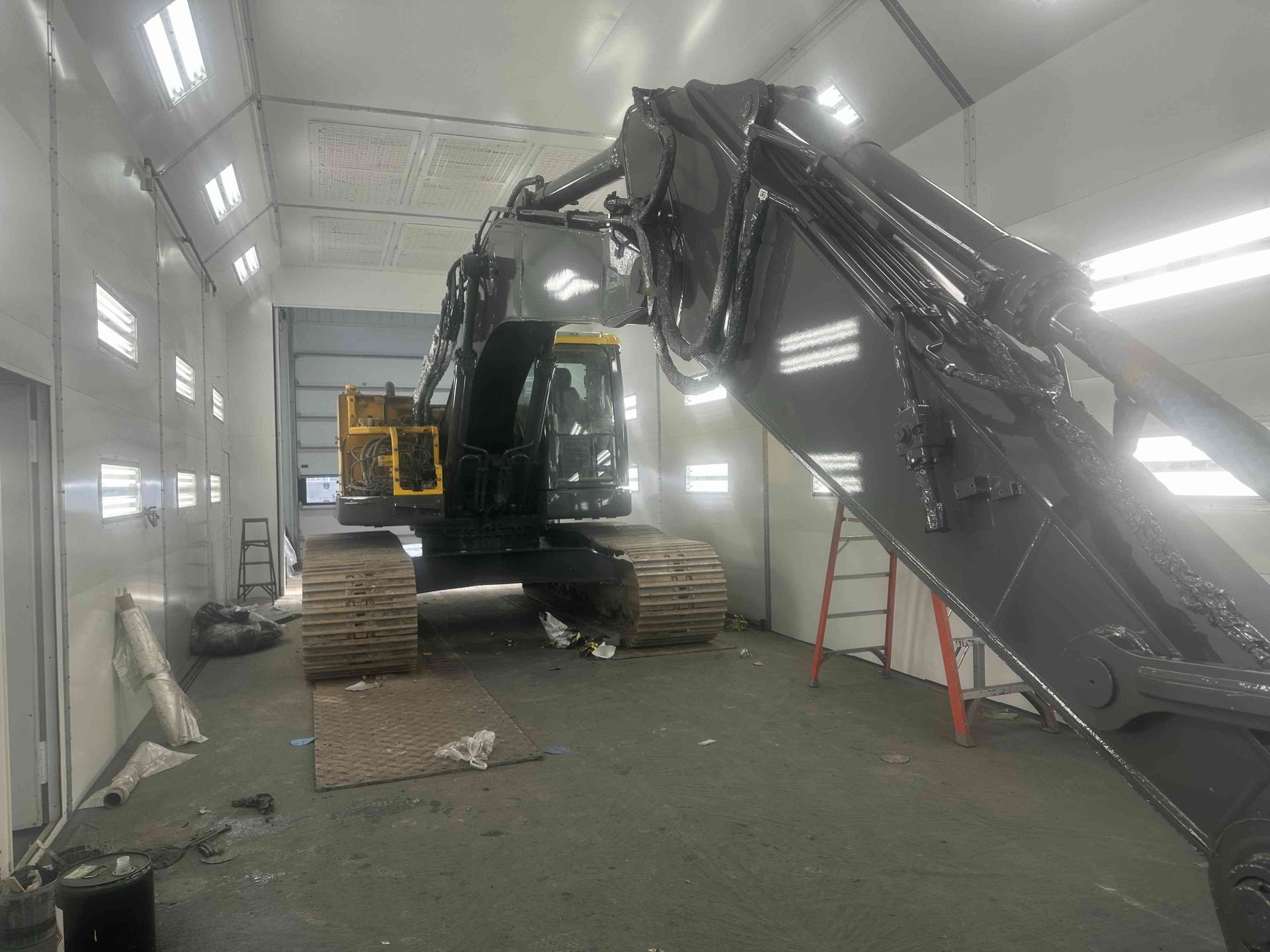 Excavator inside a white industrial spray booth, boom raised and tracks on the floor.