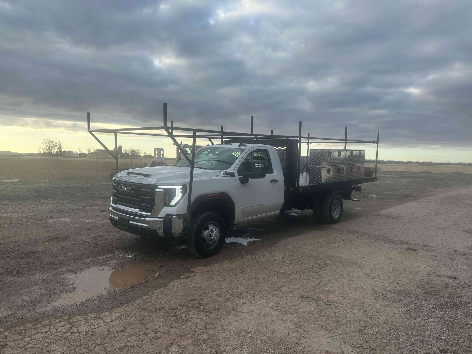 White flatbed work truck parked on a dirt road under a cloudy sky, with wet ground nearby