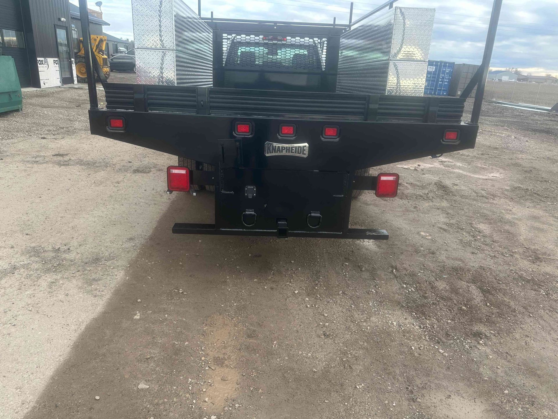 Rear view of a black flatbed trailer parked on a gravel lot, with red lights and a license plate.