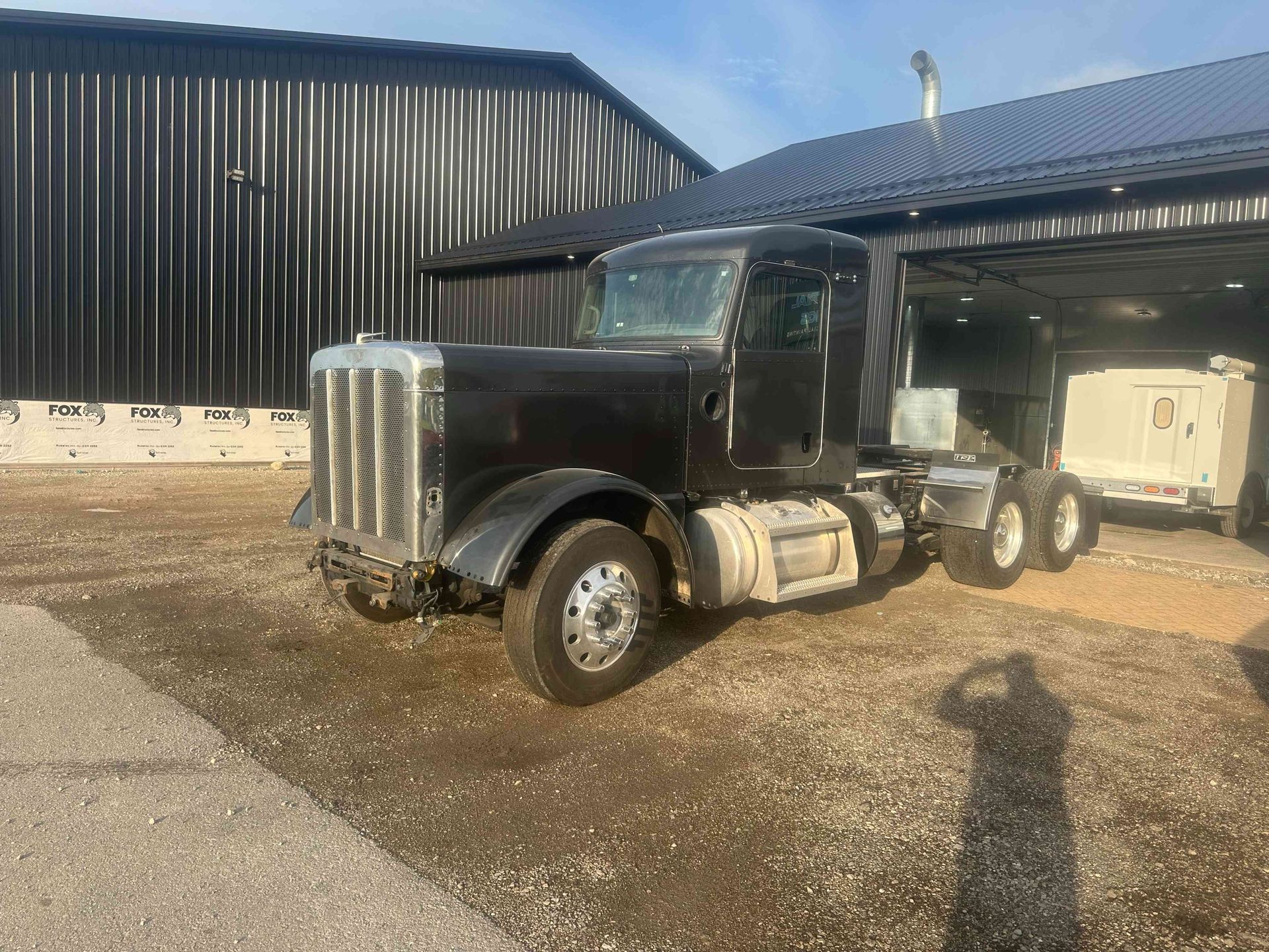 Black semi-truck parked outside a barn-like building on a gravel lot, with sunlight and open trailer space.