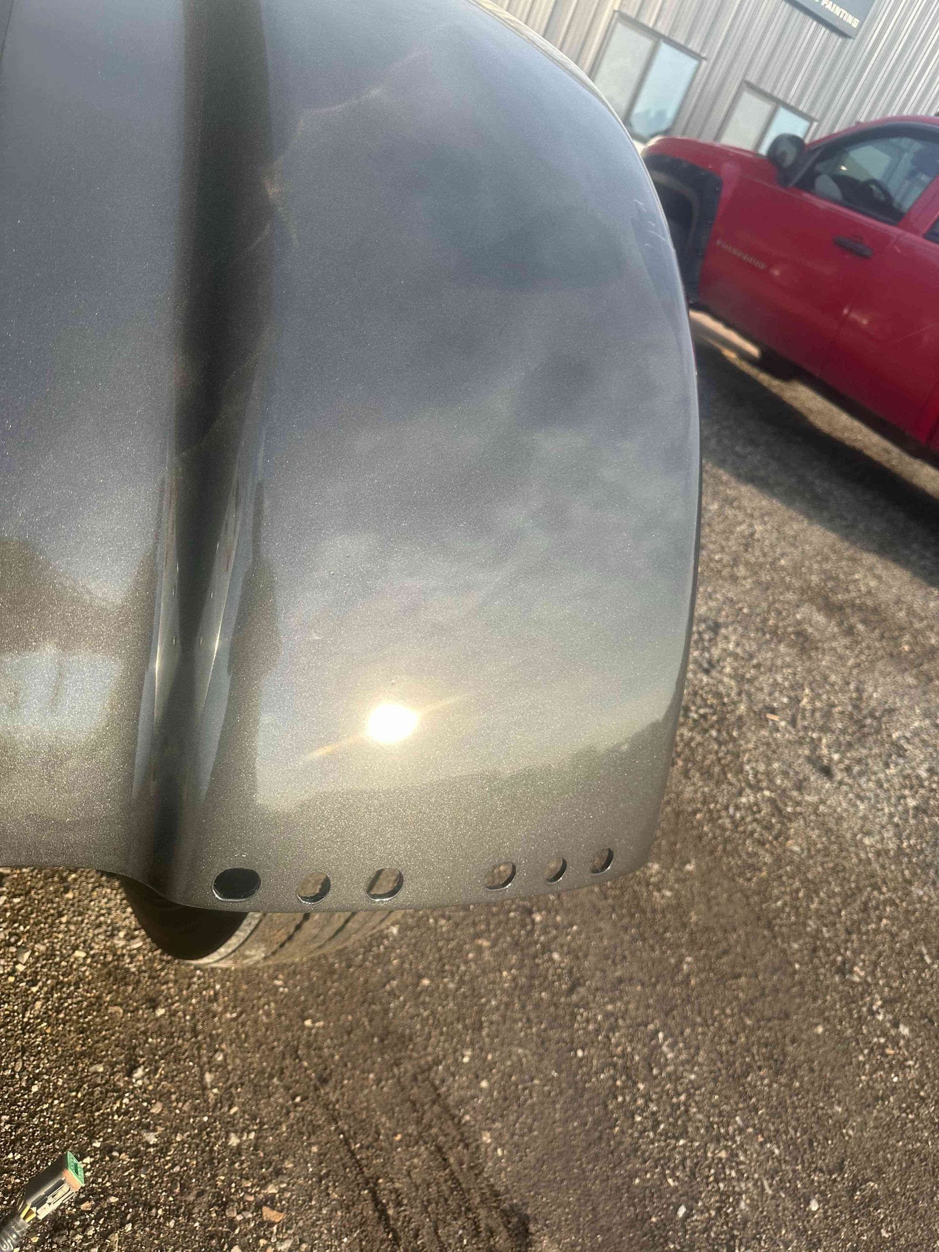 Close-up of a gray curved car part with small holes, parked on gravel near a red truck.
