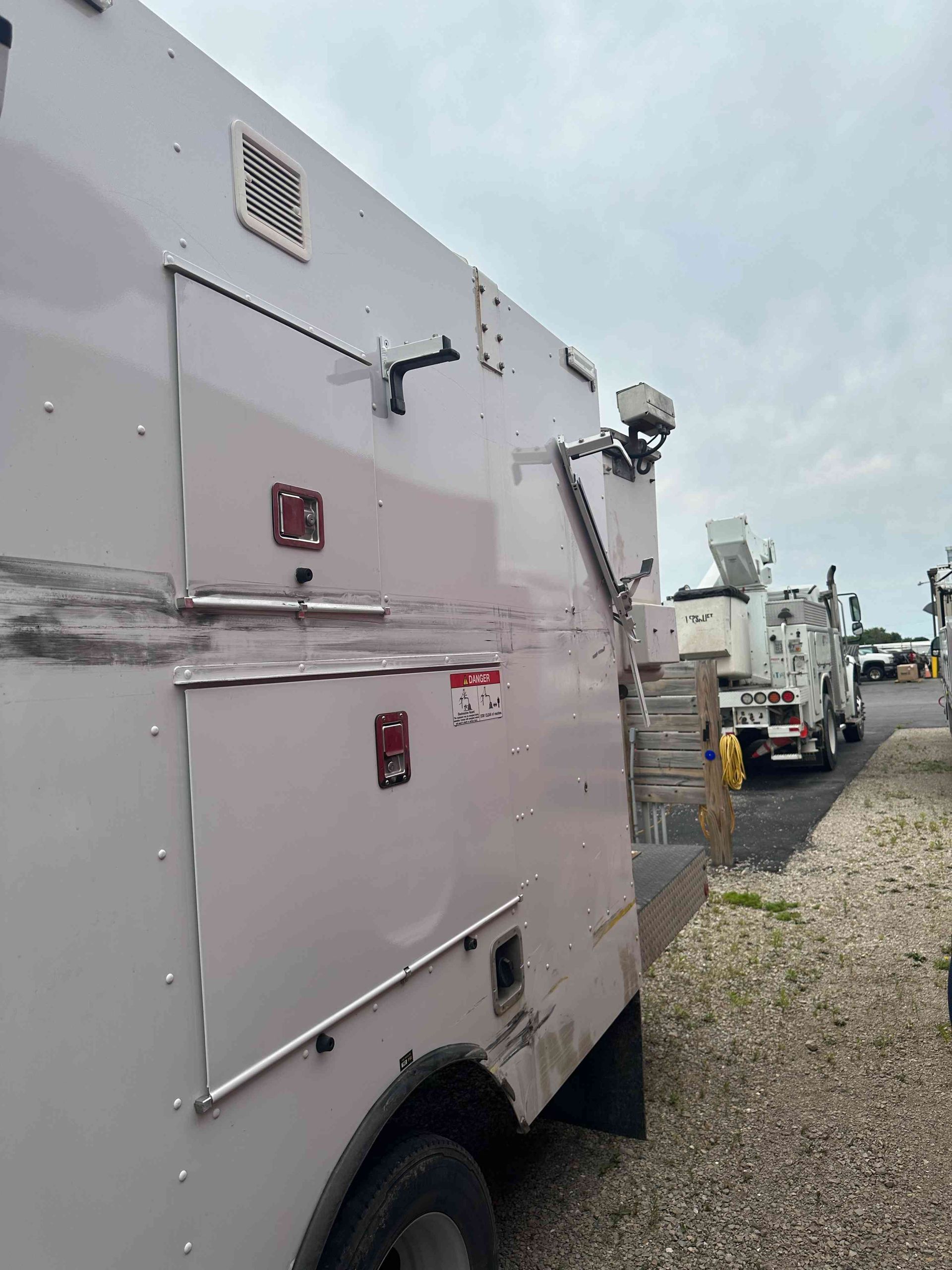 White utility truck parked on gravel beside other work vehicles under a cloudy sky