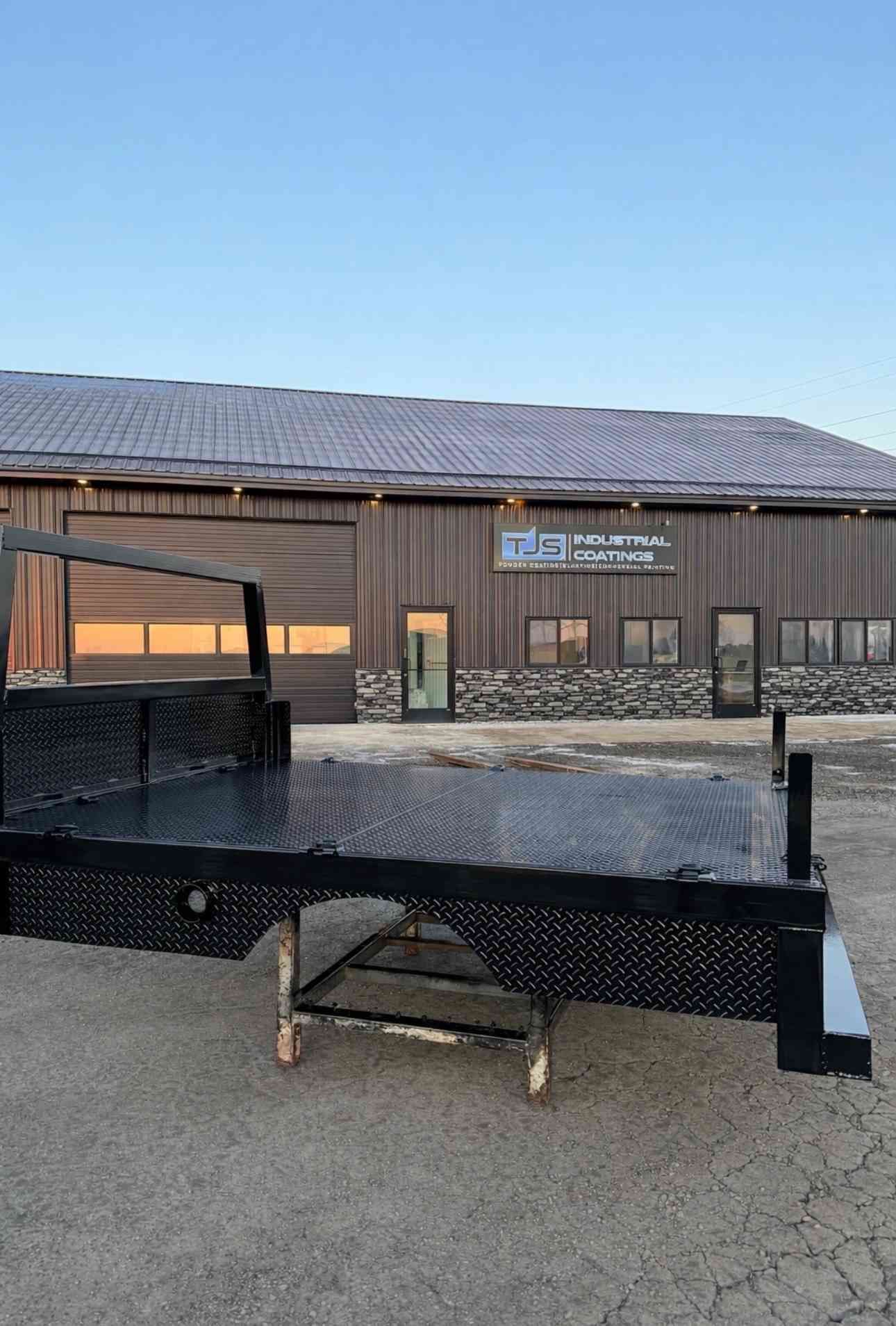 Black flatbed trailer parked in front of a metal building at dusk