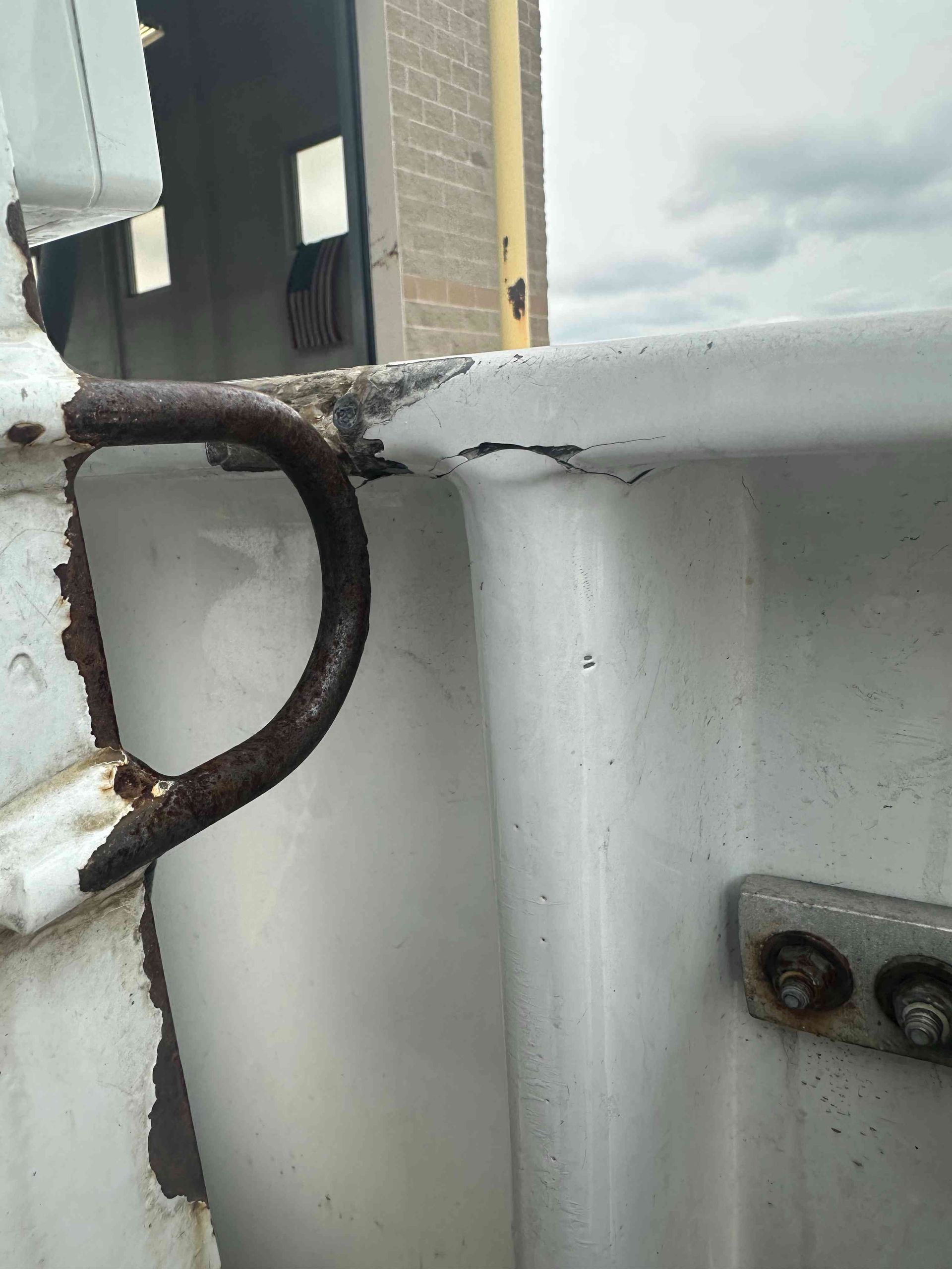 Rusty metal hook on a chipped white railing, with a building and cloudy sky in the background