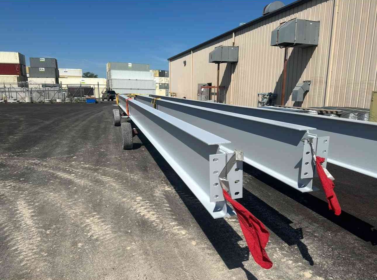 Steel bridge beams on an industrial lot beside a warehouse, with red support brackets visible.