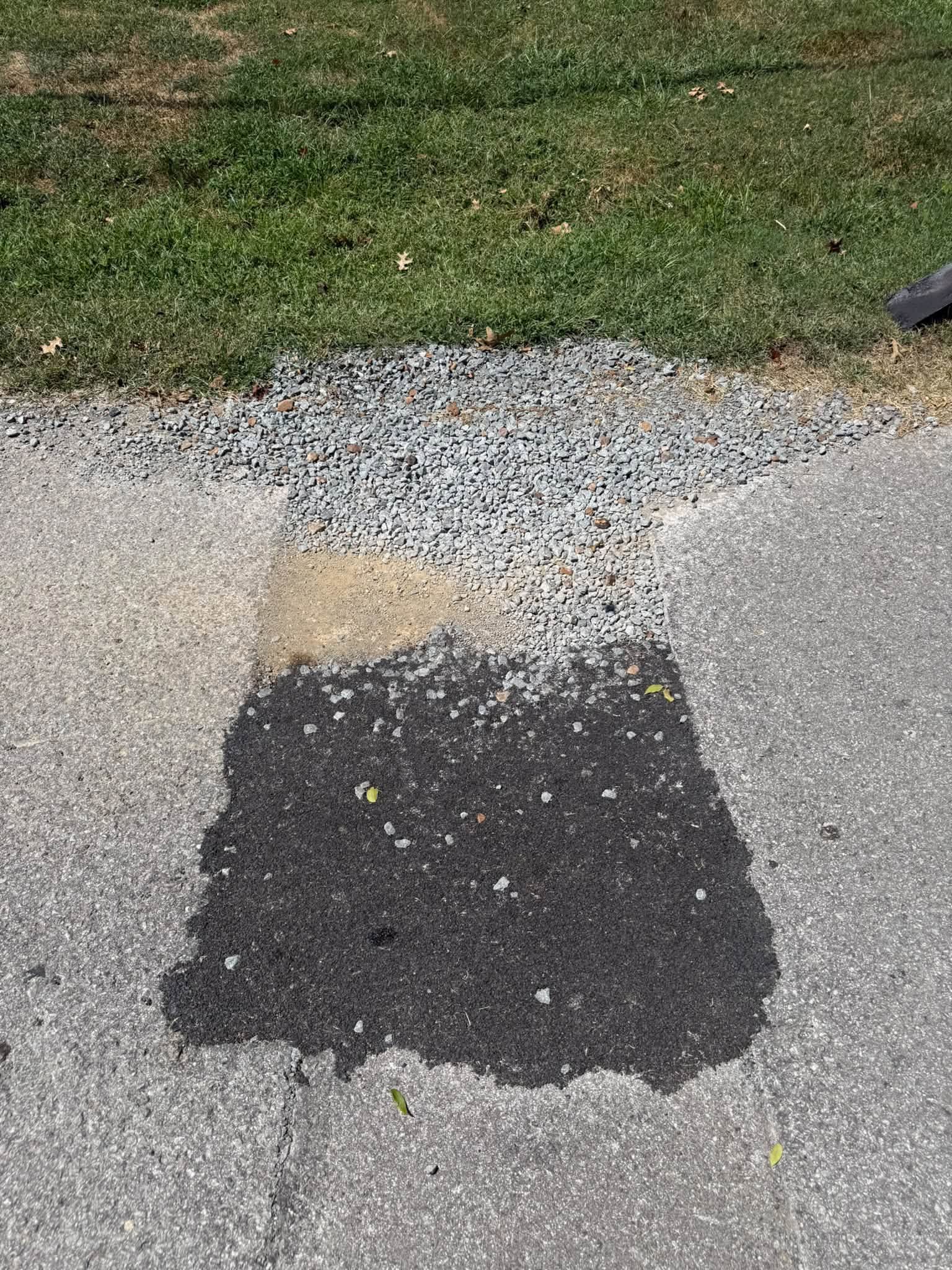 Cracked asphalt patch on a driveway edge beside grass and gravel.