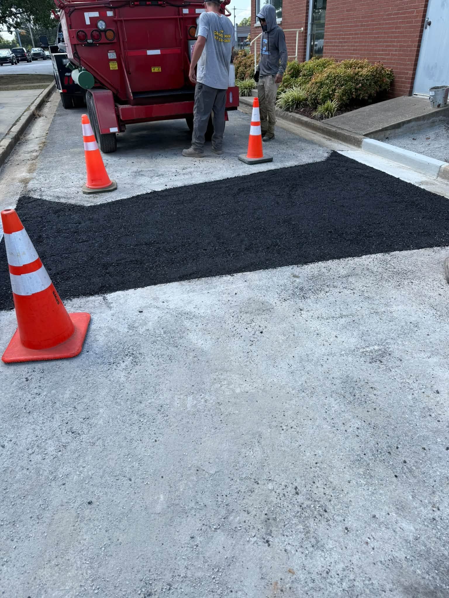 Roadwork area with fresh black asphalt patch, orange cones, and a worker beside a red truck near a driveway