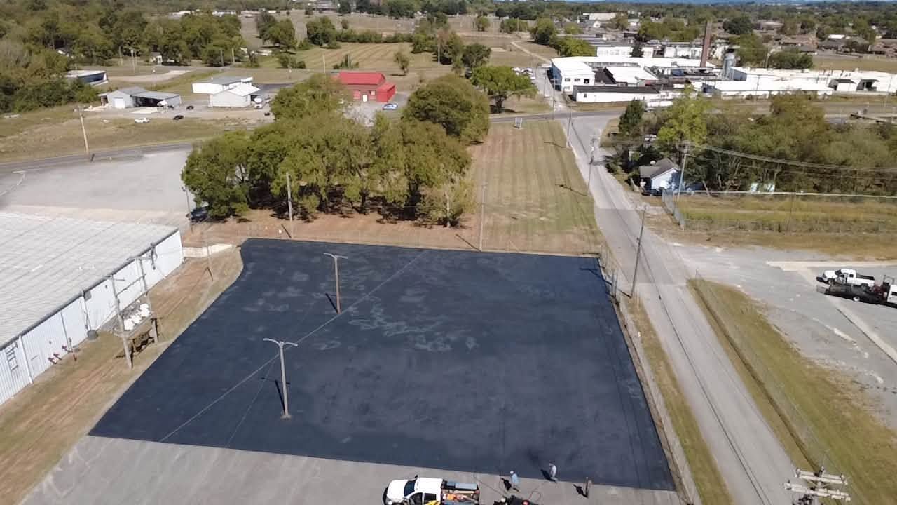 Aerial view of a large blacktopped lot beside a road, with trees and buildings in the background.