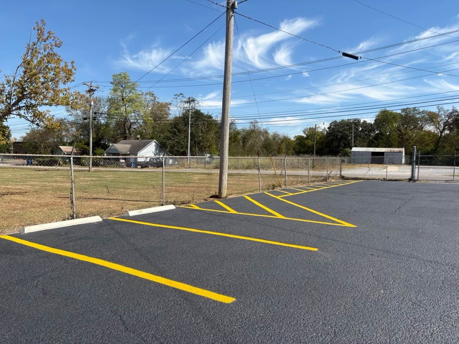 Empty paved parking lot with bright yellow diagonal lines beside a chain-link fence and trees under a blue sky