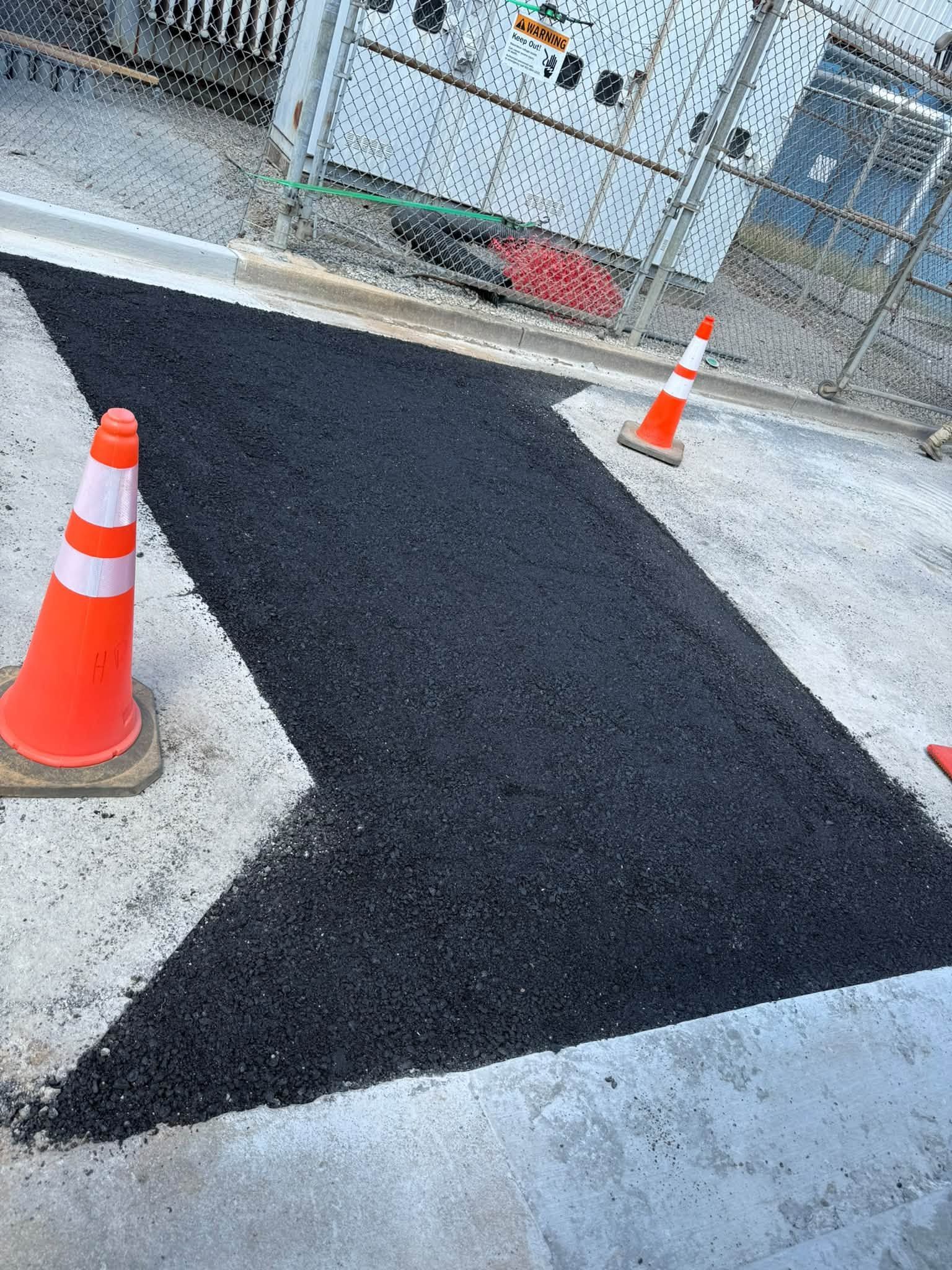 Fresh black asphalt path between orange traffic cones by a fenced gate