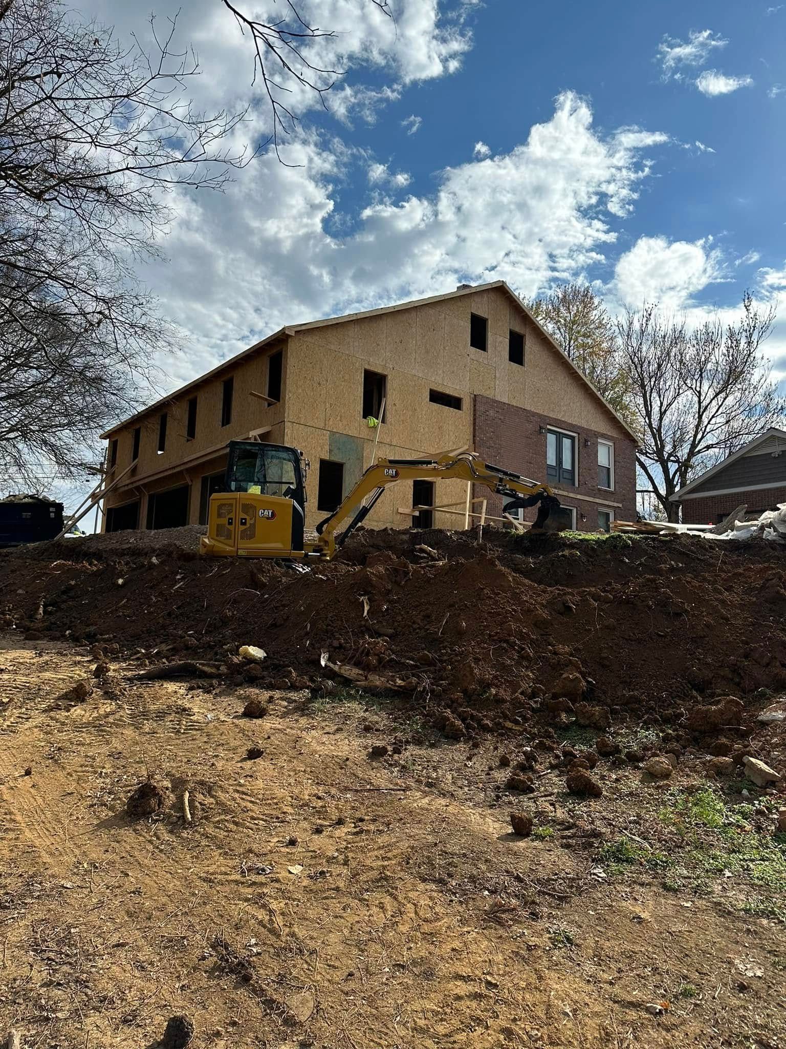 Construction of a two-story building with an excavator on a dirt mound. Blue sky with some clouds.