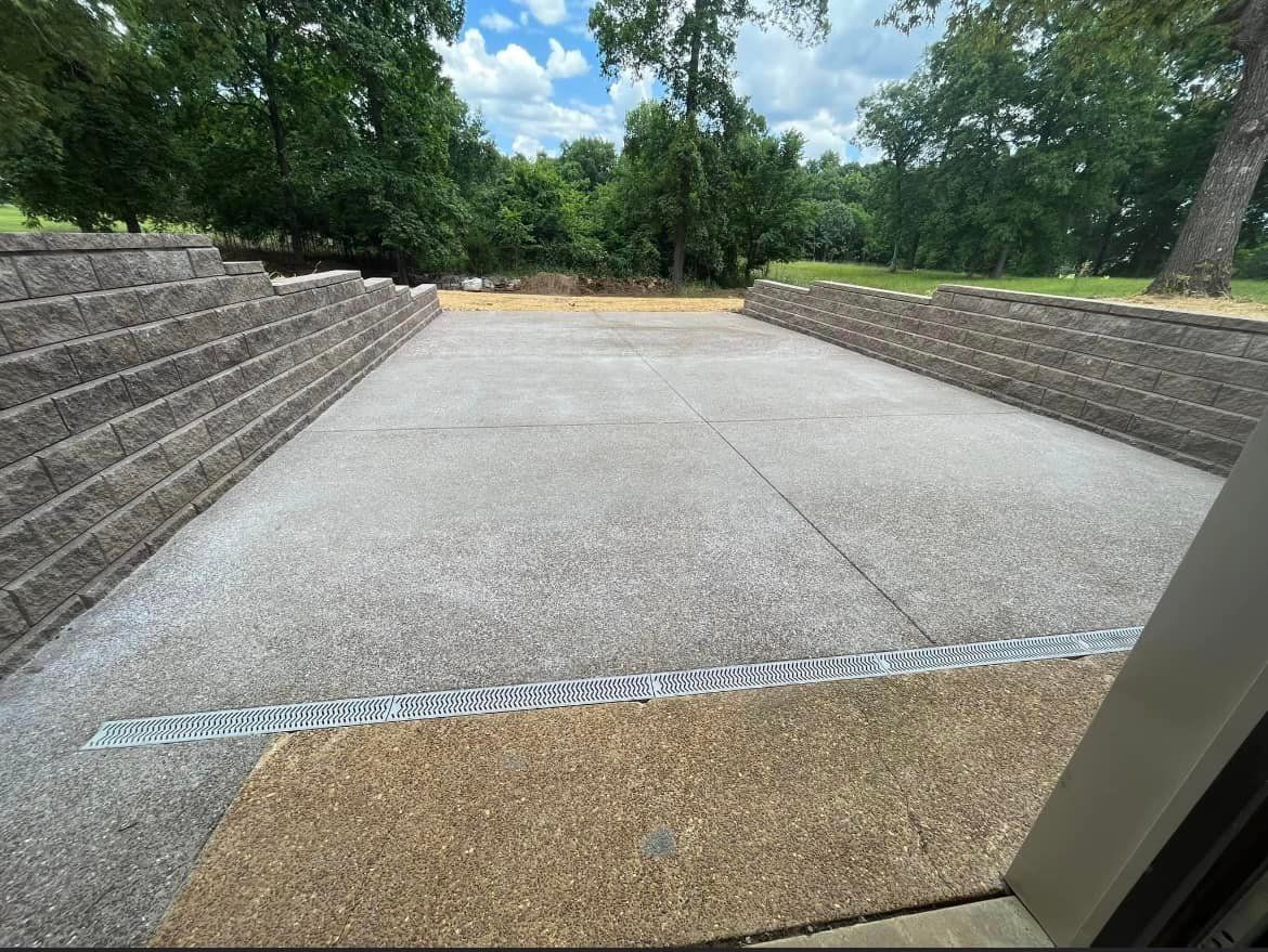 Gravel paved area with retaining walls, surrounded by trees under a blue sky.