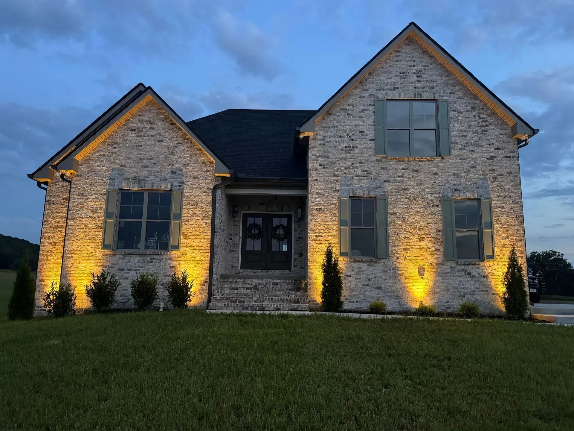 Brick house at dusk with uplighting on the facade and green shutters, set on a grassy lawn.