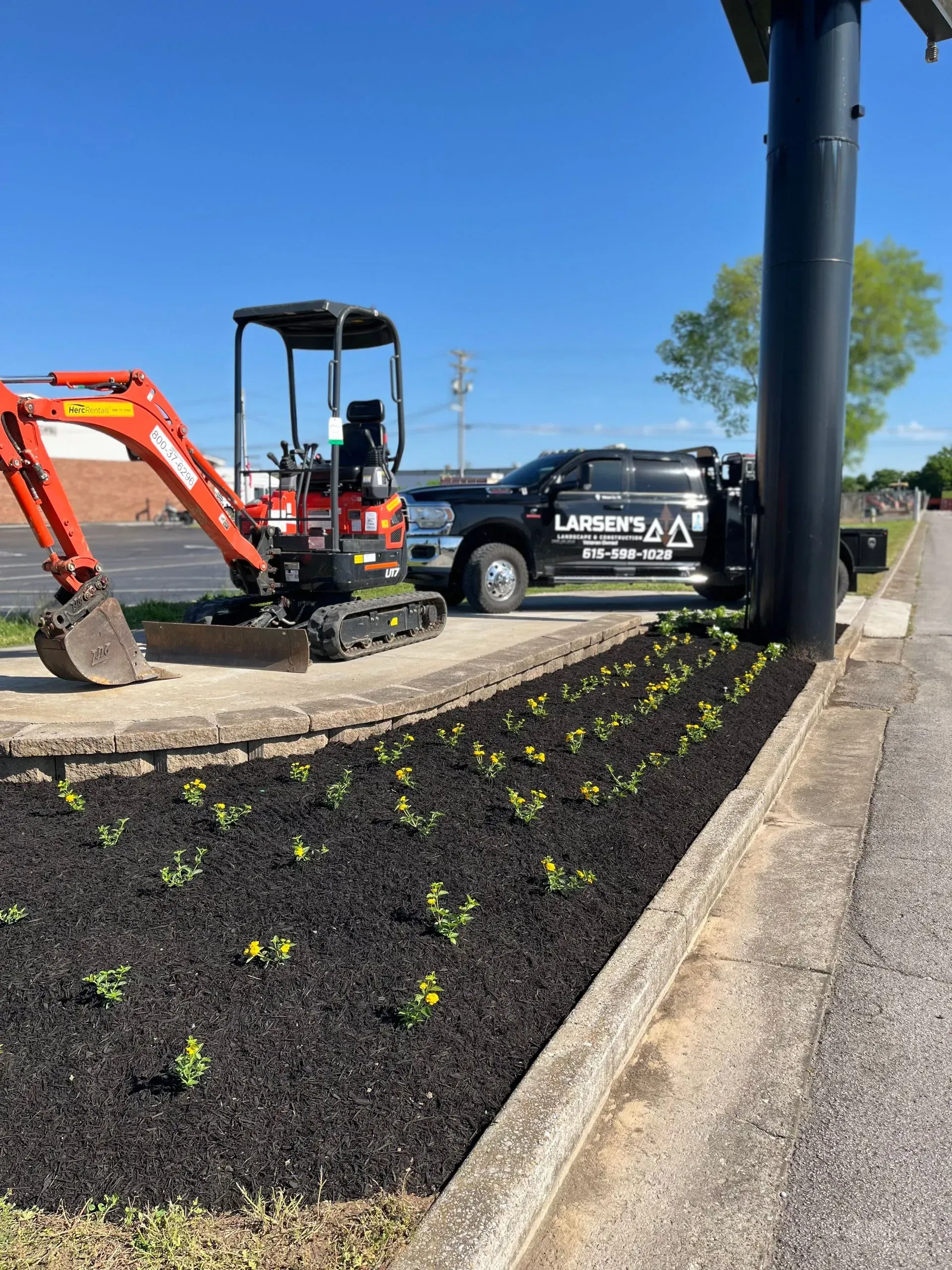 Mini-excavator and truck parked next to a newly planted garden bed filled with dark mulch and young plants.