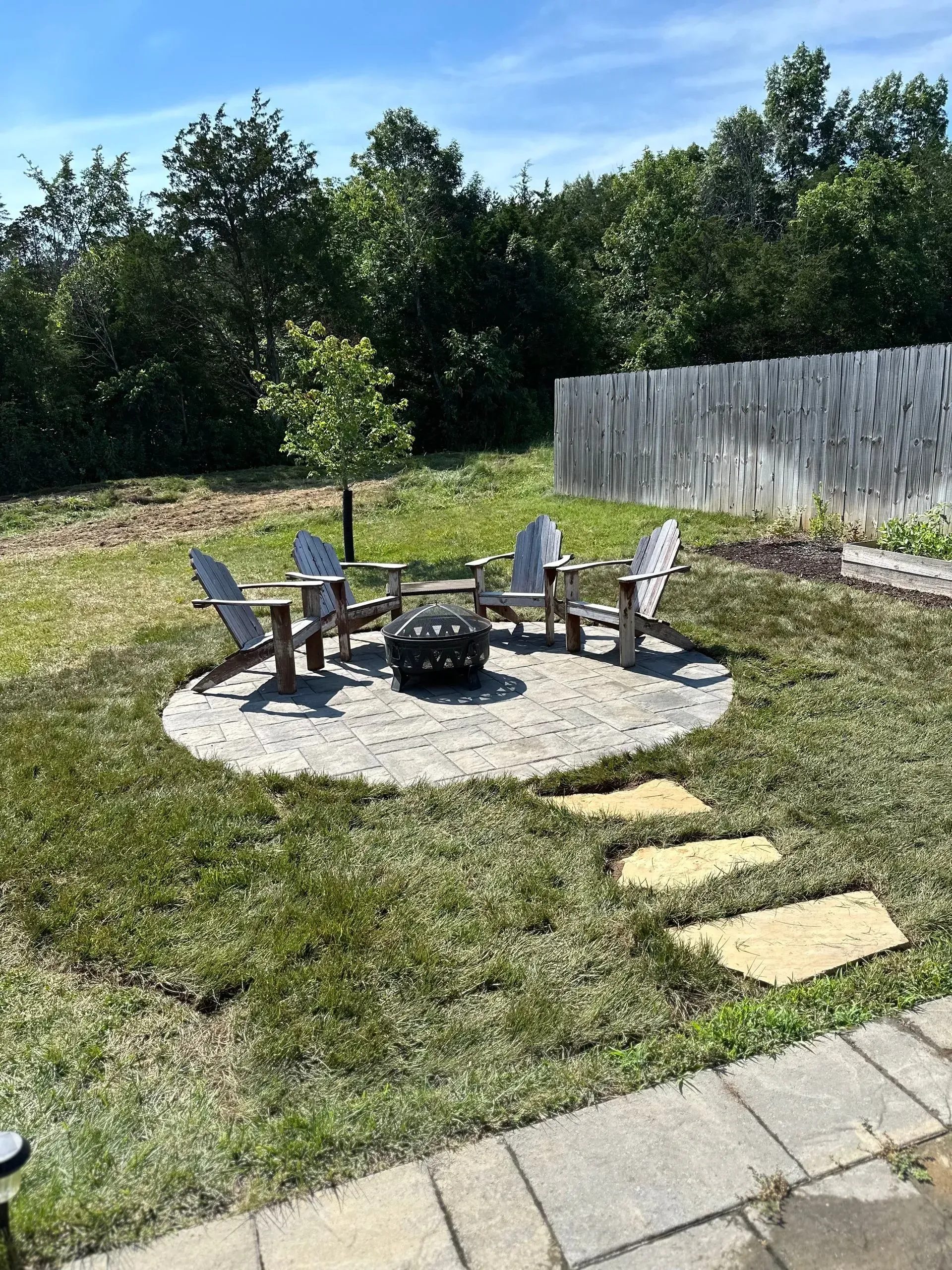 Backyard with a fire pit surrounded by Adirondack chairs on a stone patio. Yellow stepping stones lead to the pit.