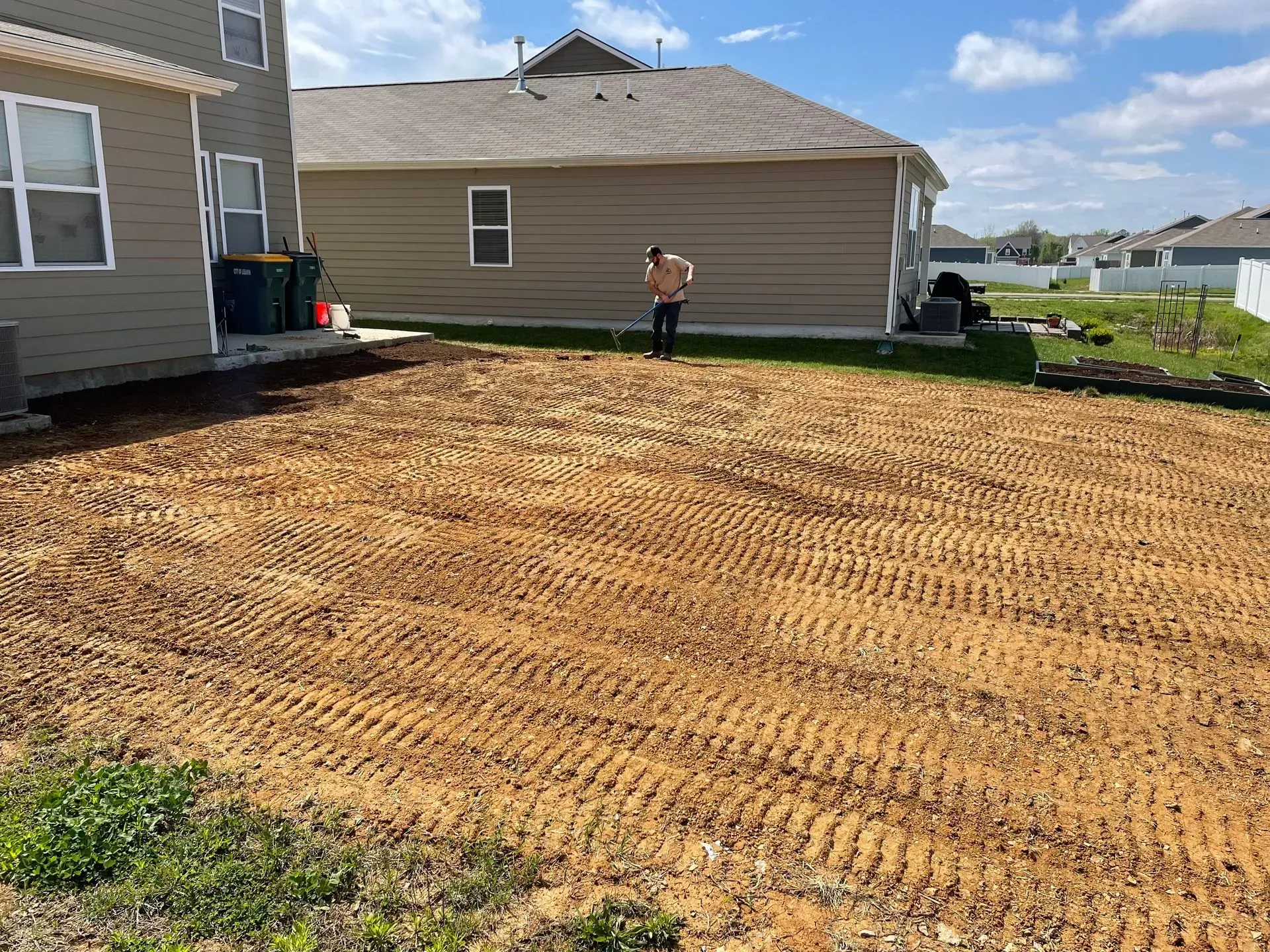 A person standing in a freshly tilled, brown dirt yard with two houses in the background on a sunny day.