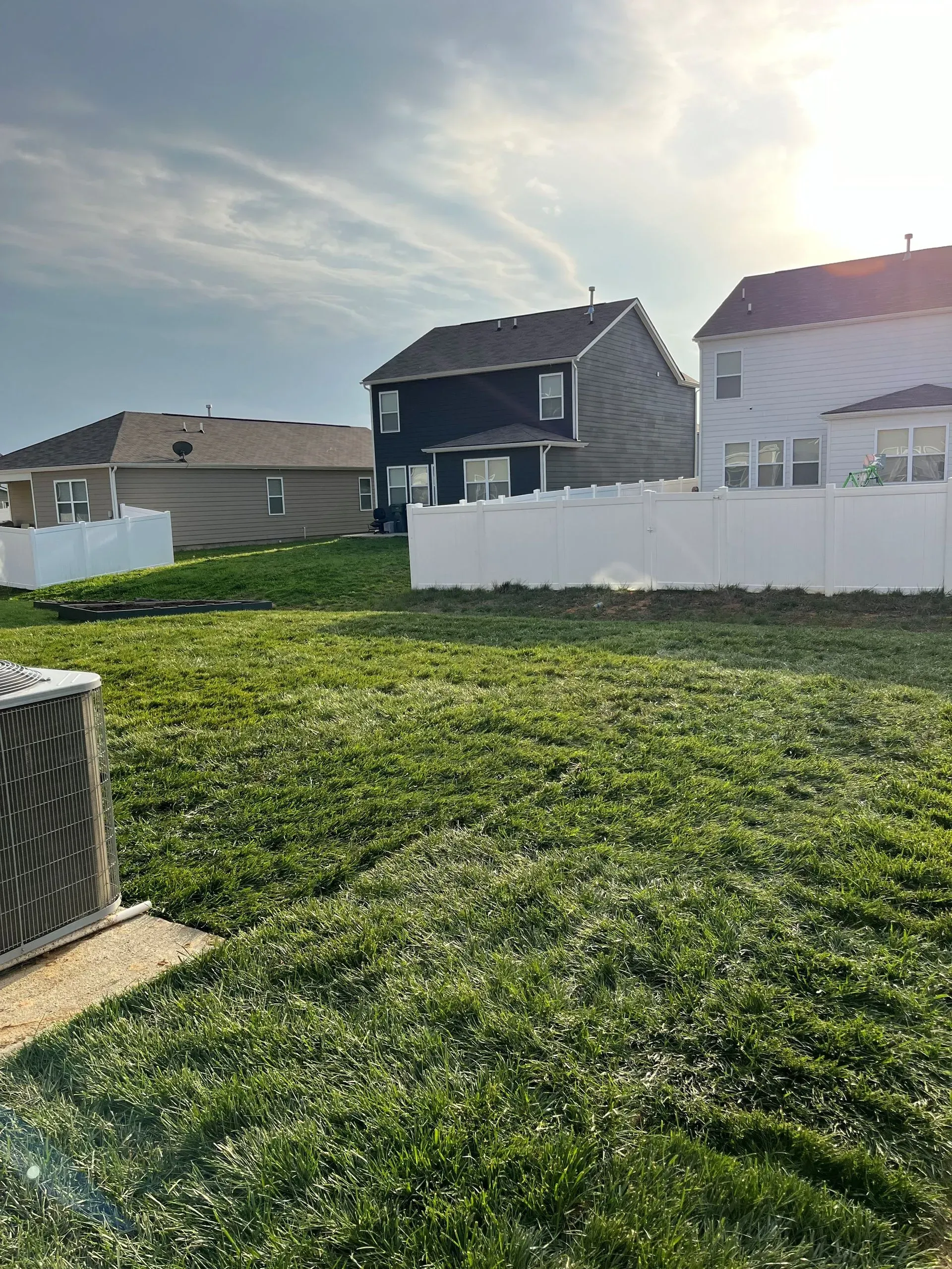 Backyard with a white fence, green grass, and houses under a cloudy sky.