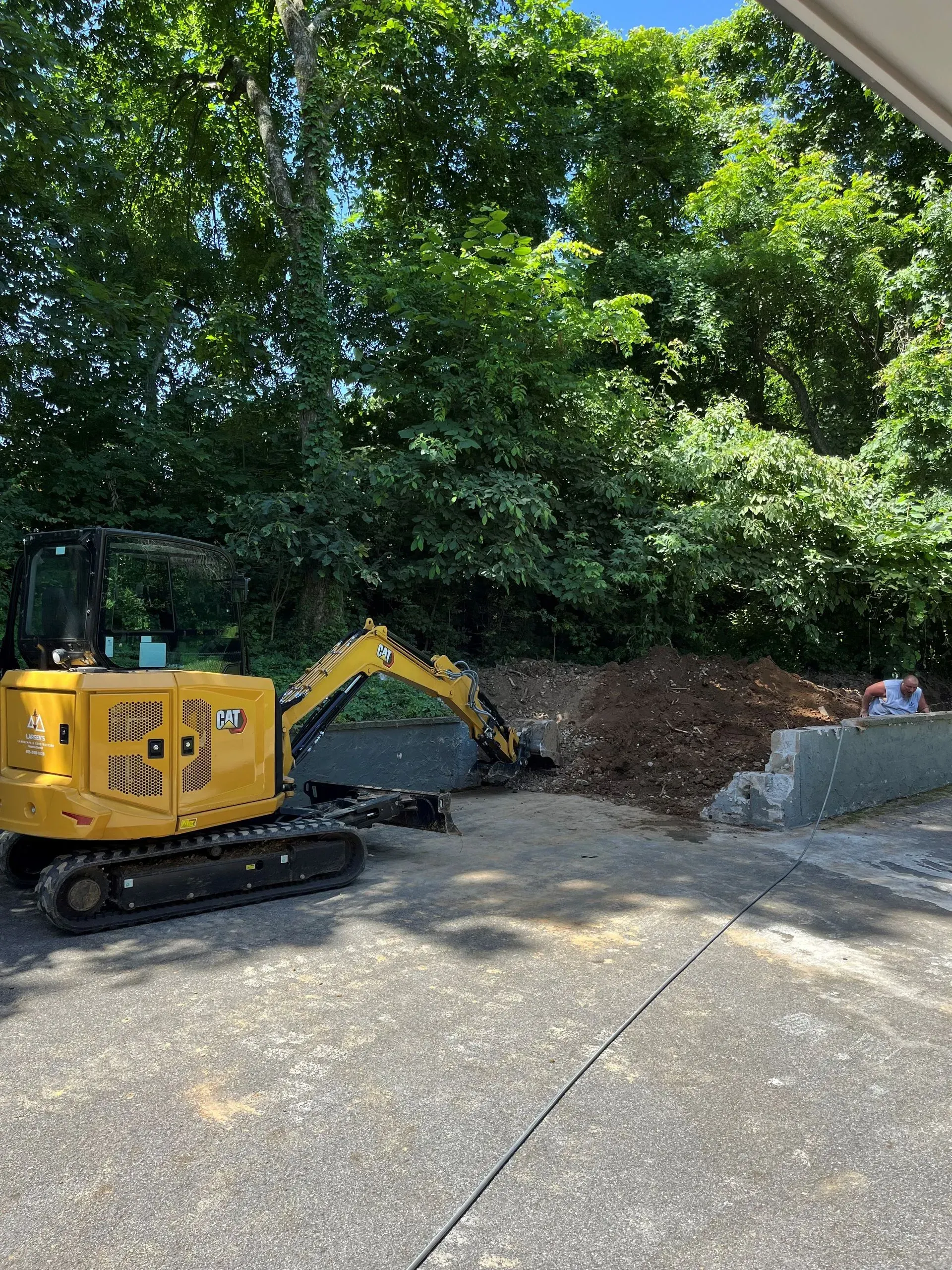 Yellow excavator working on a pile of dirt next to a retaining wall, trees in the background.