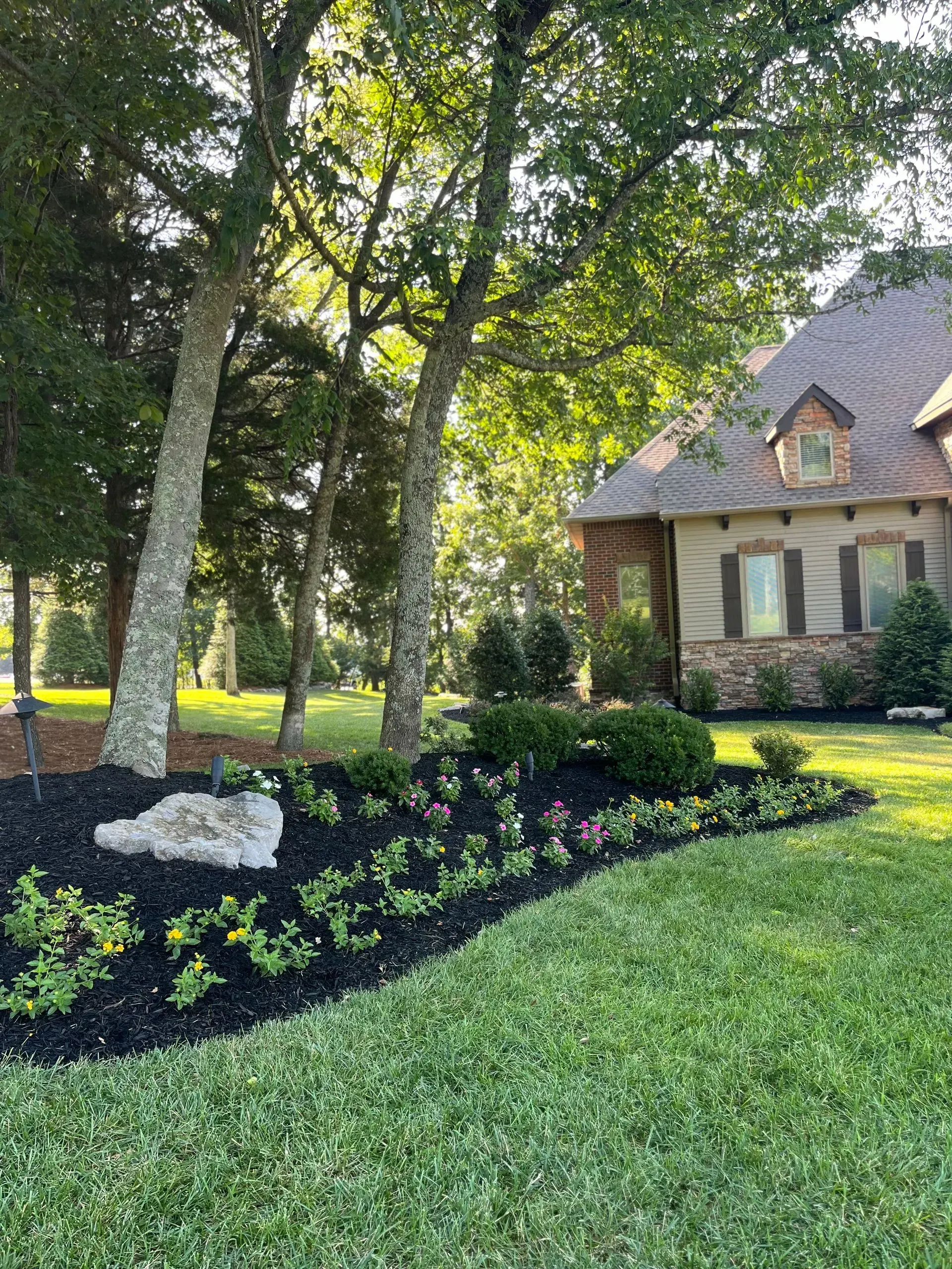 Landscaped front yard with flower bed, mulch, and house under trees.