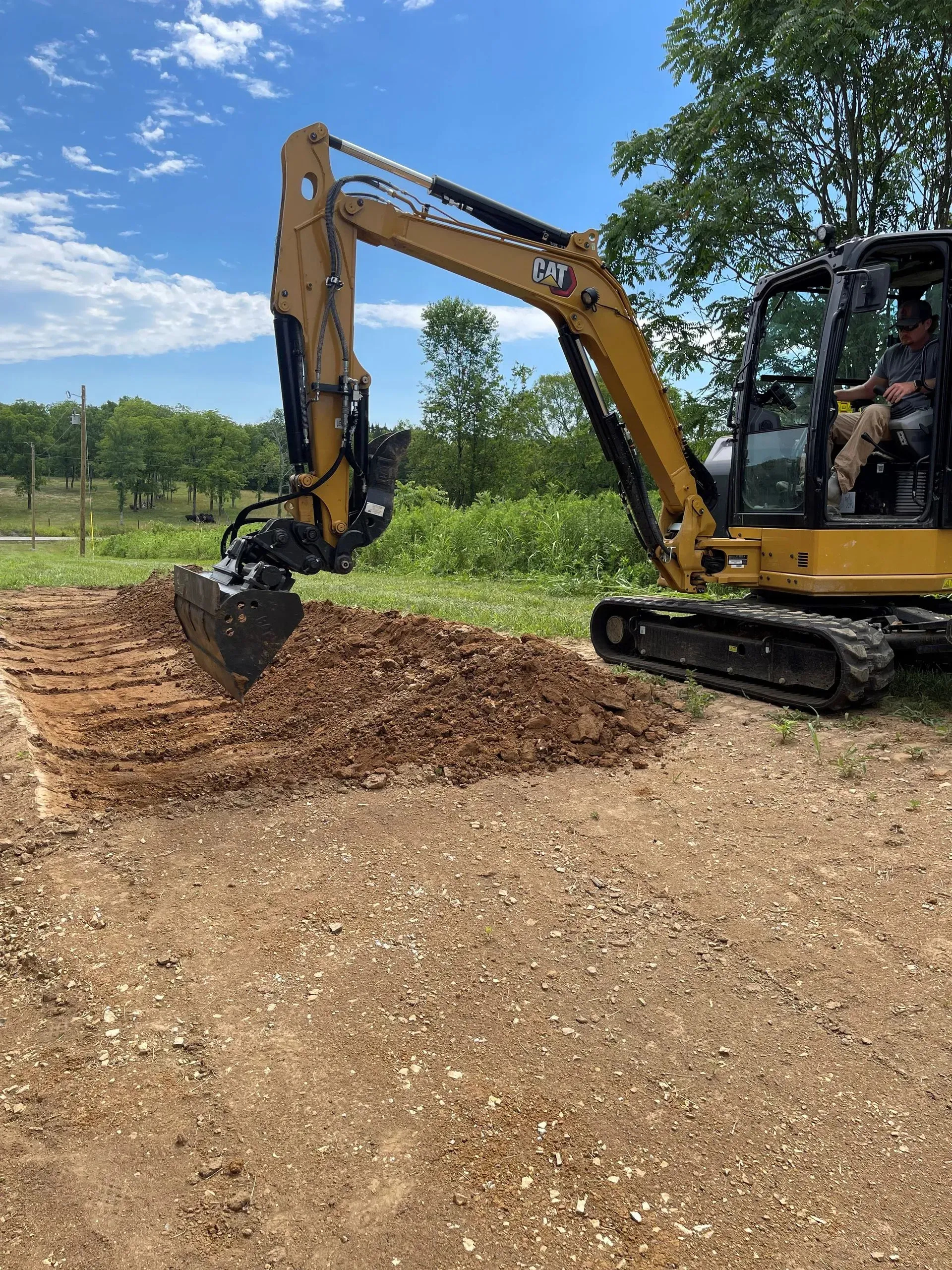 Yellow excavator digging in dirt, operator visible inside cab, sunny outdoors.