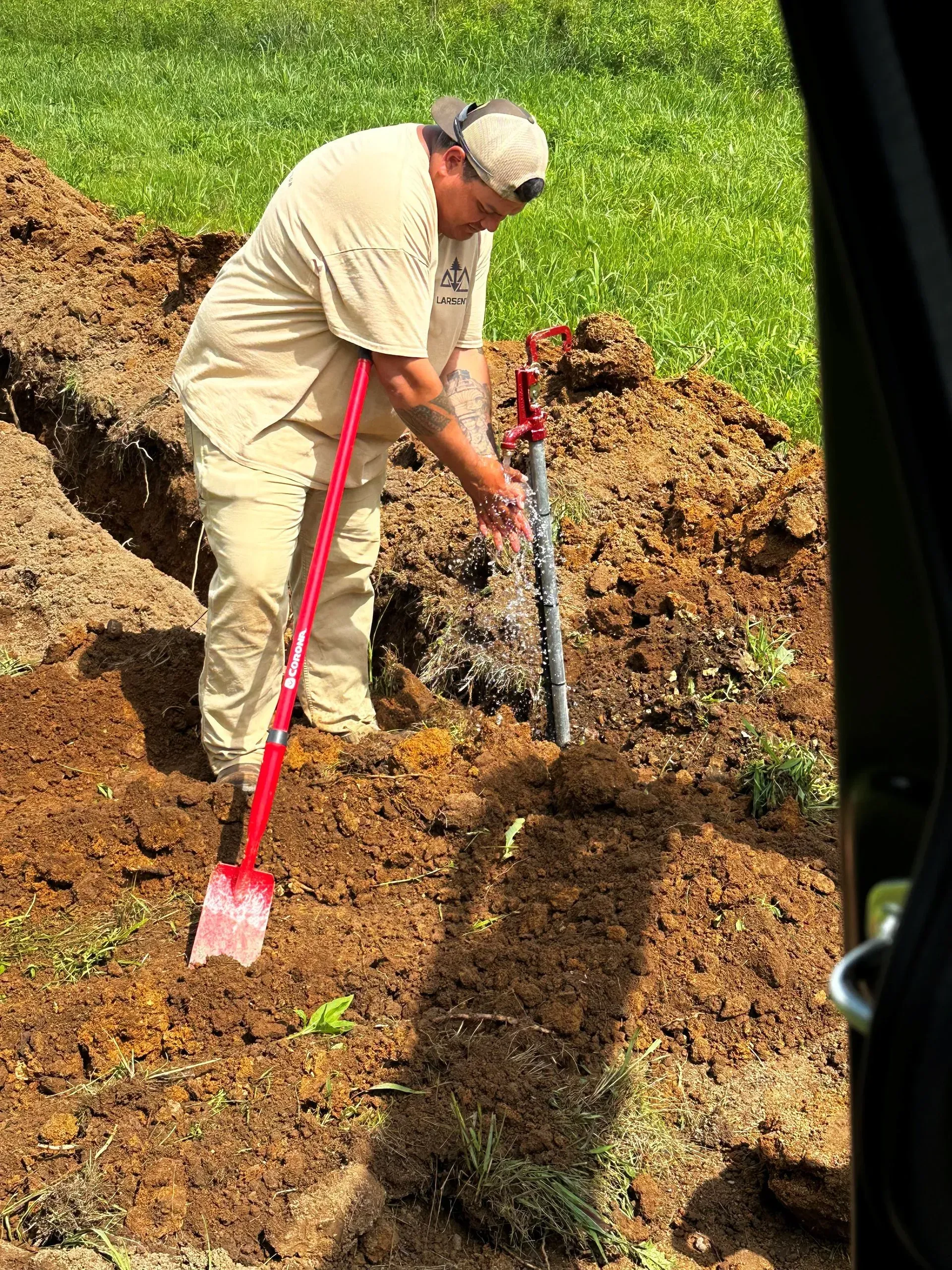 Man digging in dirt with a shovel next to a pipe, wearing tan clothes and a hat, outdoors.