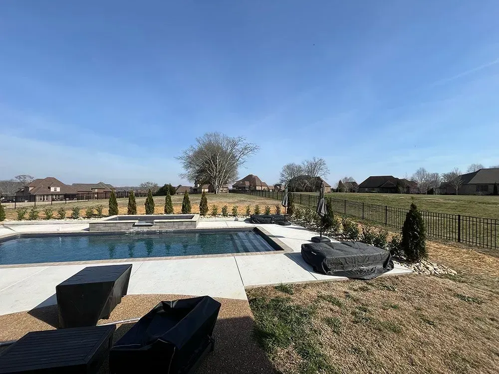 A backyard pool with patio furniture, surrounded by grass and trees, under a blue sky.